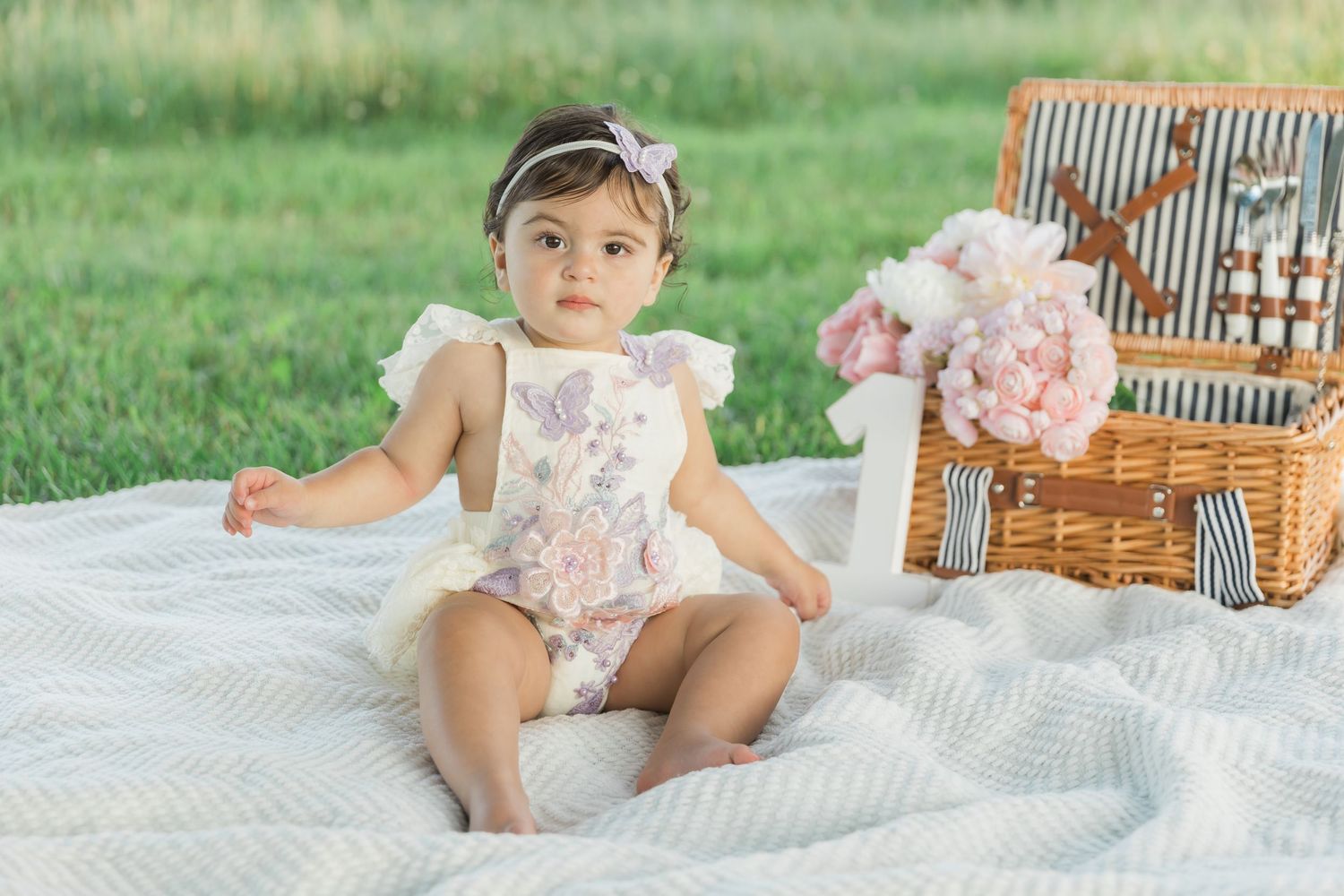 Floral romper wearing baby sits on white blanket next to picnic basket with pink flowers outdoors on grass.