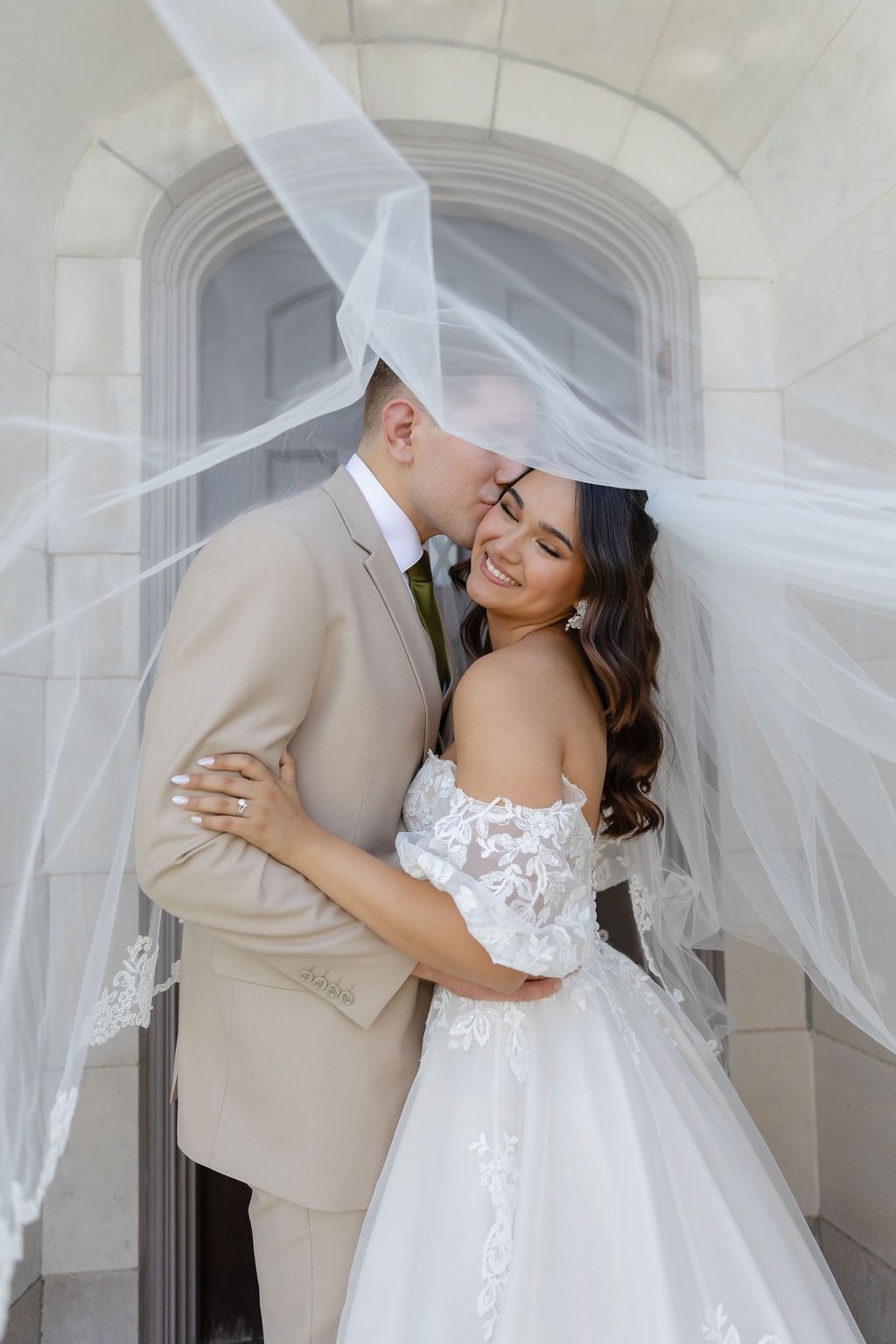 Romantic wedding photos under flowing white veil as couple embraces in elegant outdoor setting.