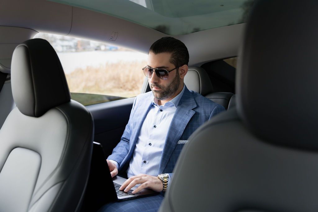 Businessman in blue suit sitting in back of luxury car while working with laptop and wearing sunglasses.
