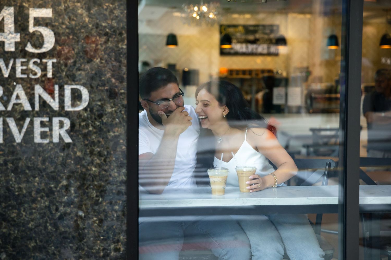 Couple sharing tender moment viewed through cafe window with street number visible.