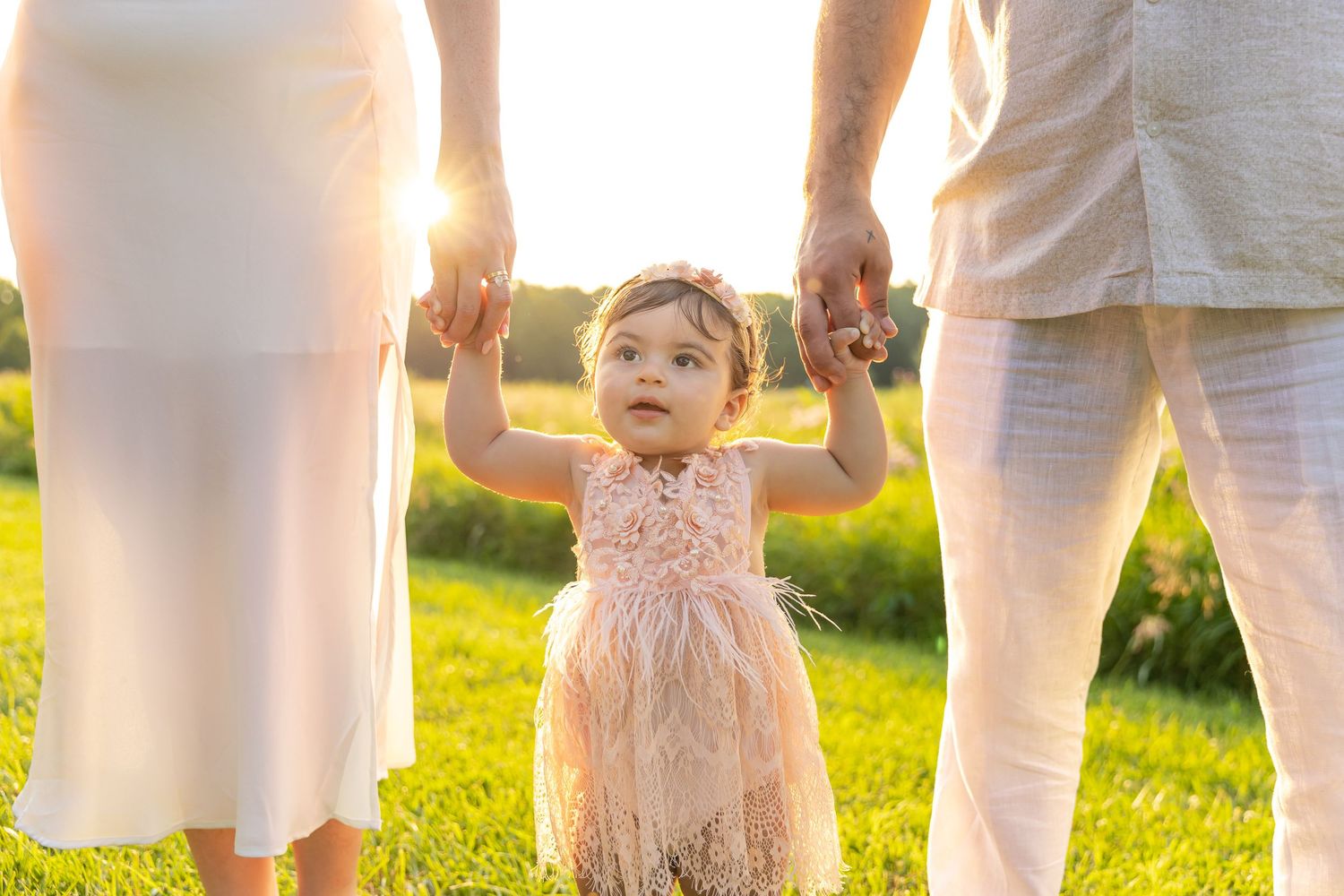 A small child in a pale dress stands holding hands with two people in white clothing at sunset in a grassy field.