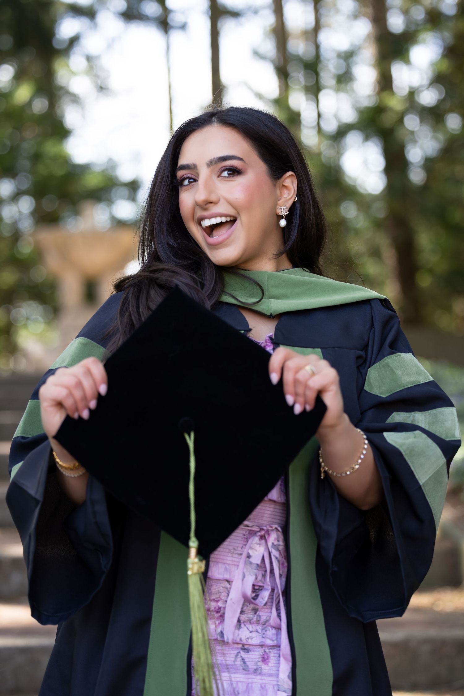 A graduate in academic regalia shows excitement while holding their graduation cap outdoors.