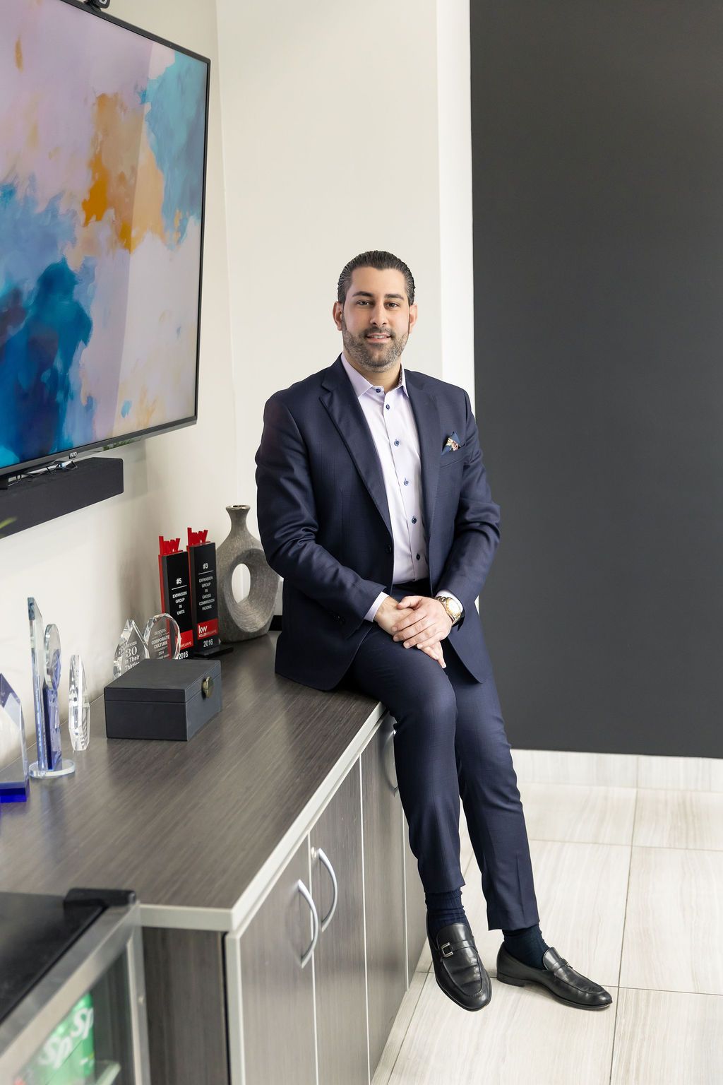 Professional in navy suit sits casually on office cabinet beside colorful abstract wall art and drink bottles.