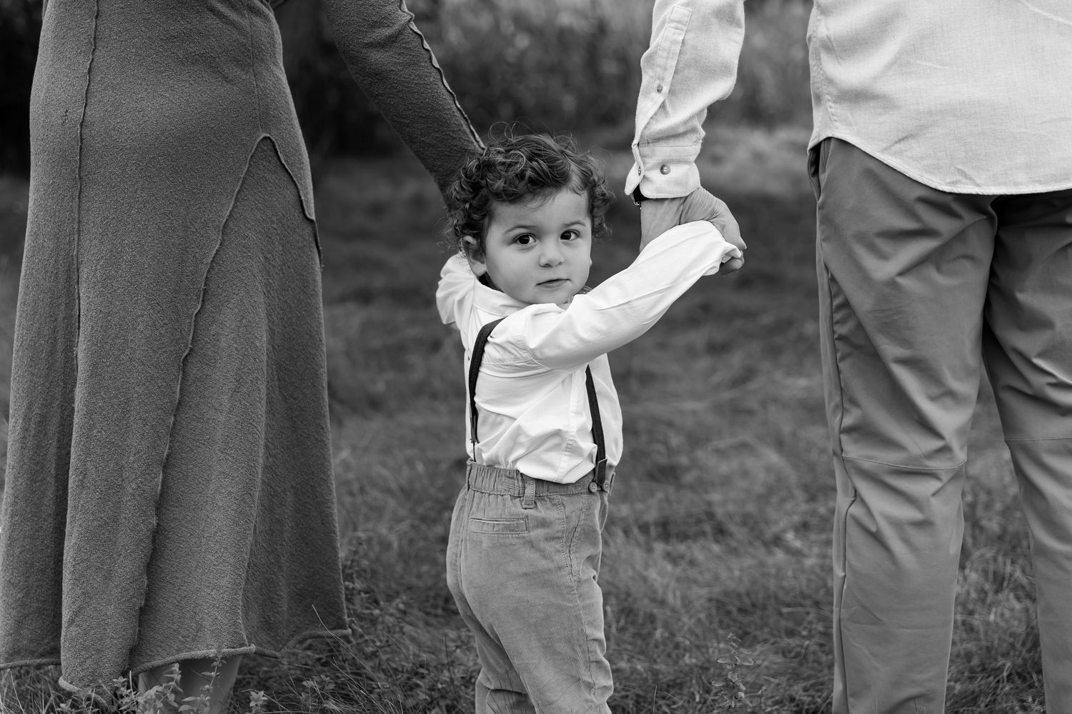 Black and white photo of a small child in suspenders holding hands with adults outdoors.