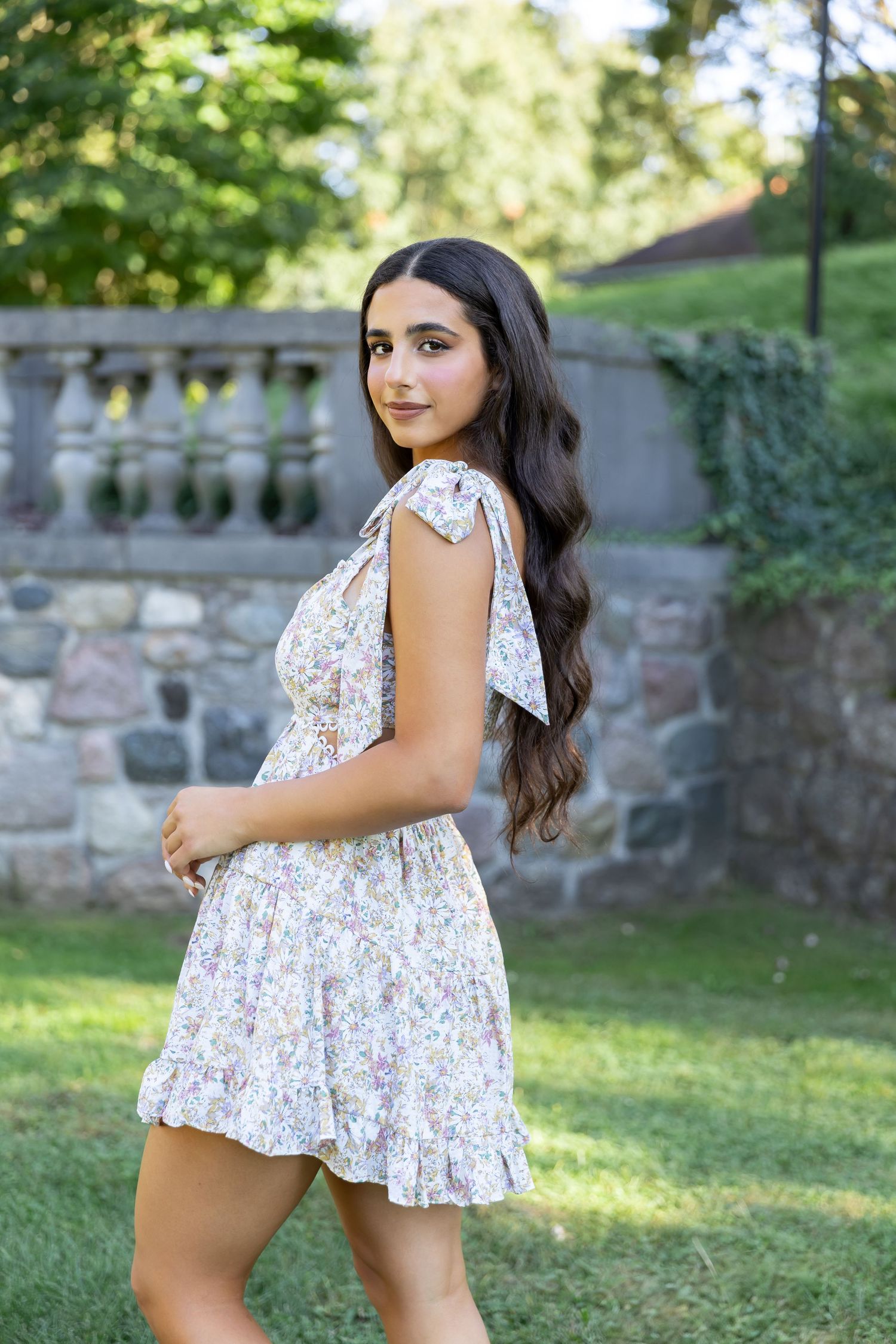 Woman in floral dress posing in garden setting with stone wall and greenery.