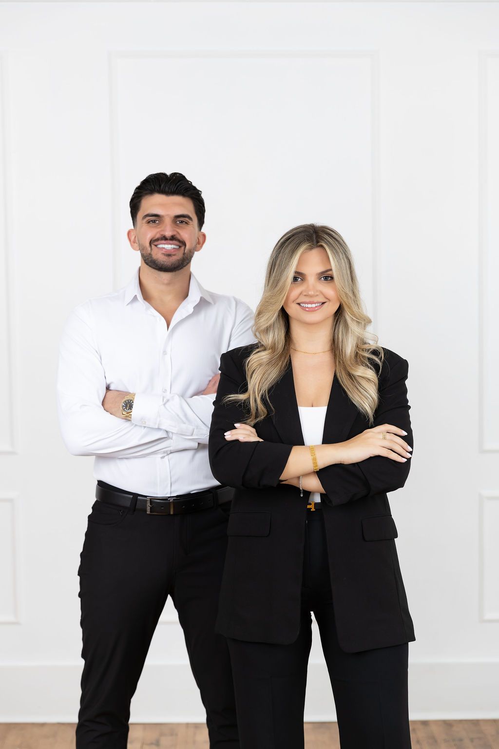 Professional business colleagues in formal attire stand confidently with arms crossed against white wall.