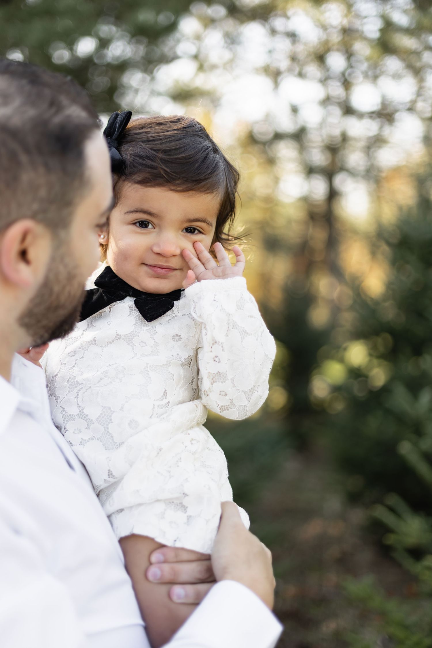 Parent in white shirt holding adorable child in white lace dress against blurred nature background.