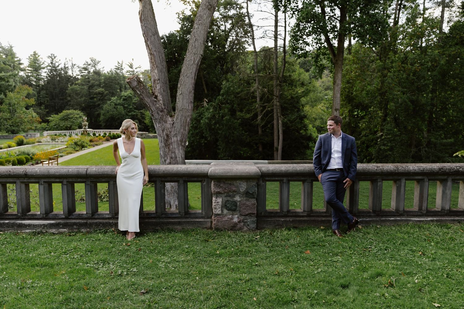 Bride in white dress and groom in suit standing separated on garden terrace.