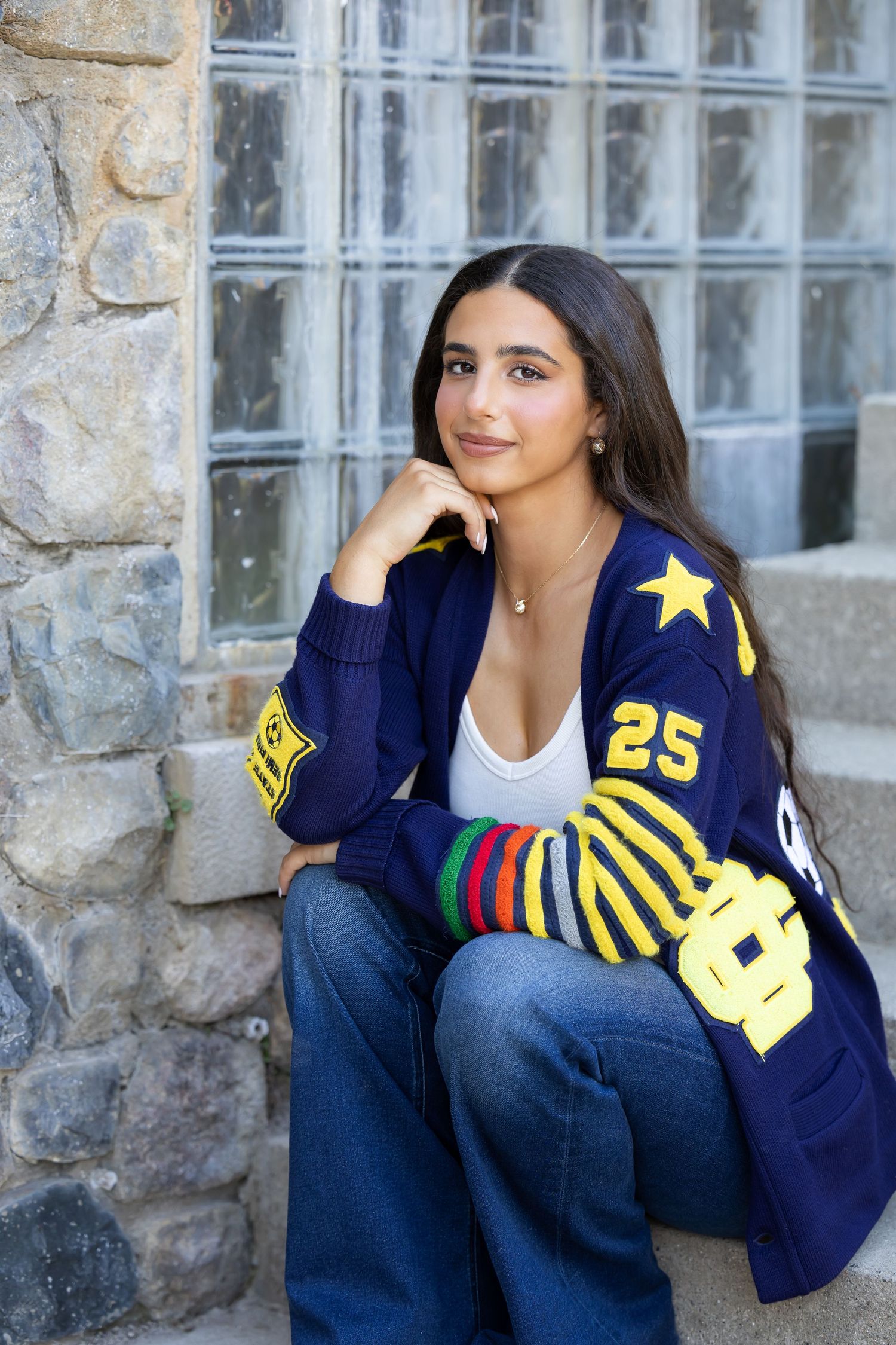 Person wearing varsity letterman jacket with number 25 poses against stone wall.