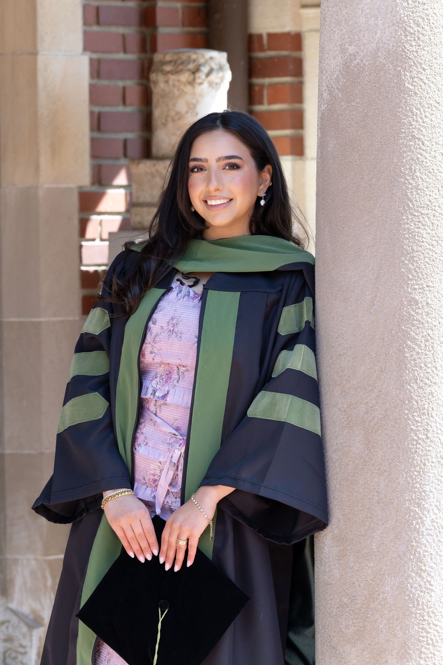 A graduate in academic regalia smiles while standing against a brick building exterior.