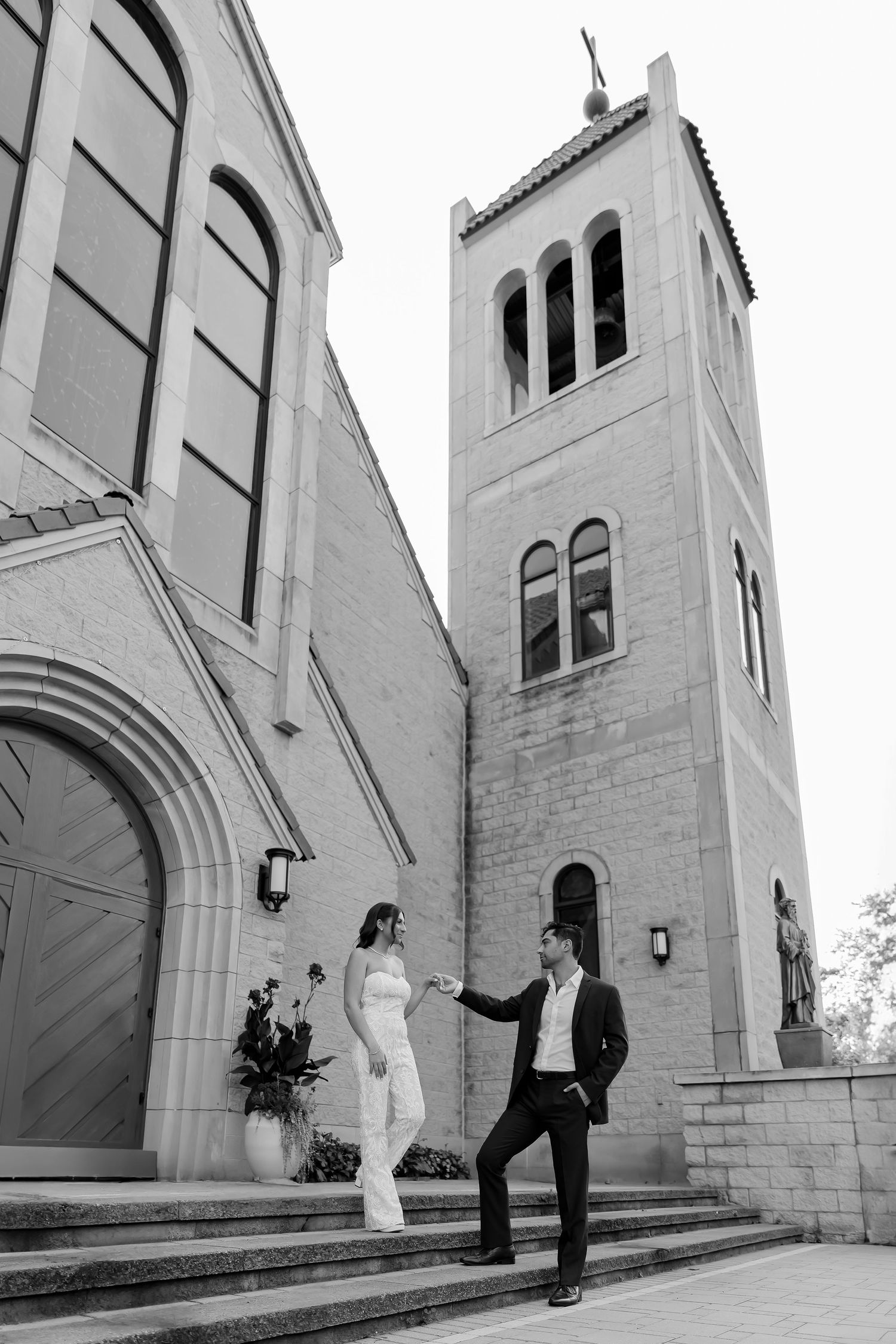 A couple stands on church steps with a white stone bell tower and arched doorway in the background.