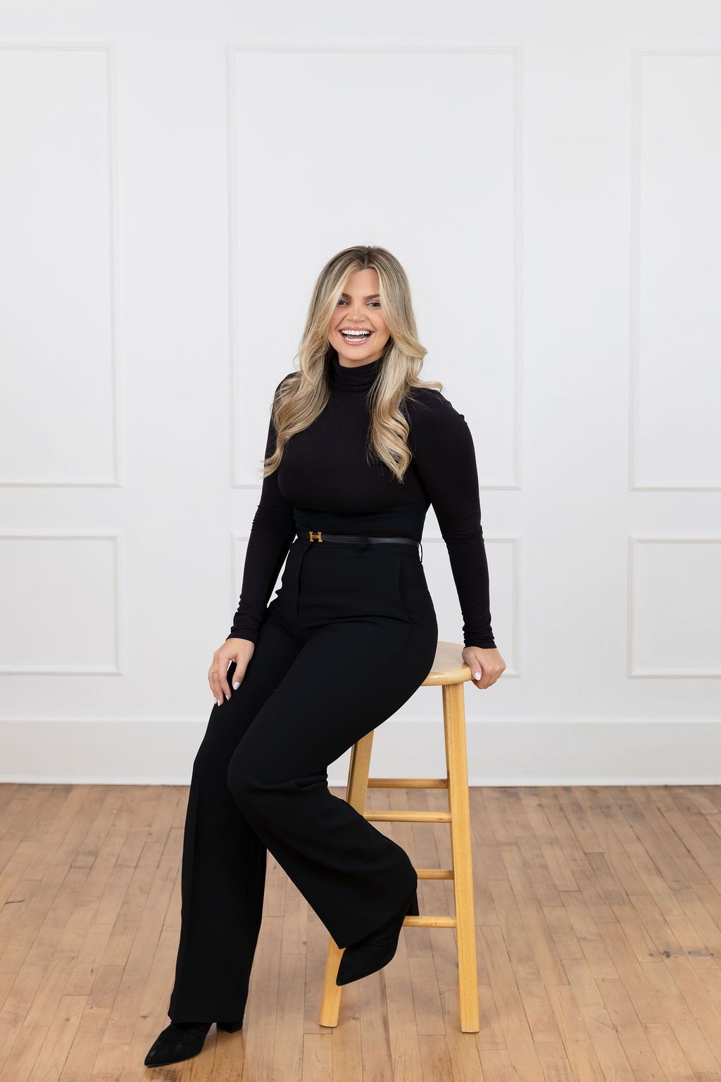 A stylish all-black ensemble featuring turtleneck and wide-leg pants paired with a belted waist while seated on wooden stool.