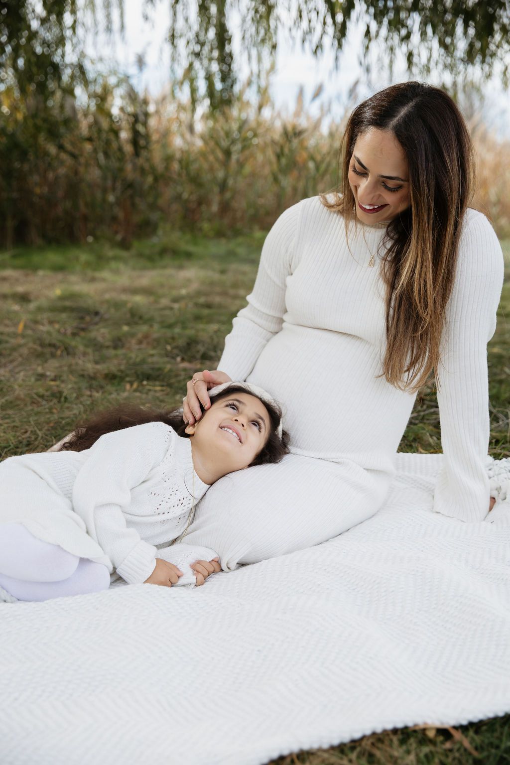 A mother and child dressed in white lay on a blanket outdoors with natural greenery in the background.