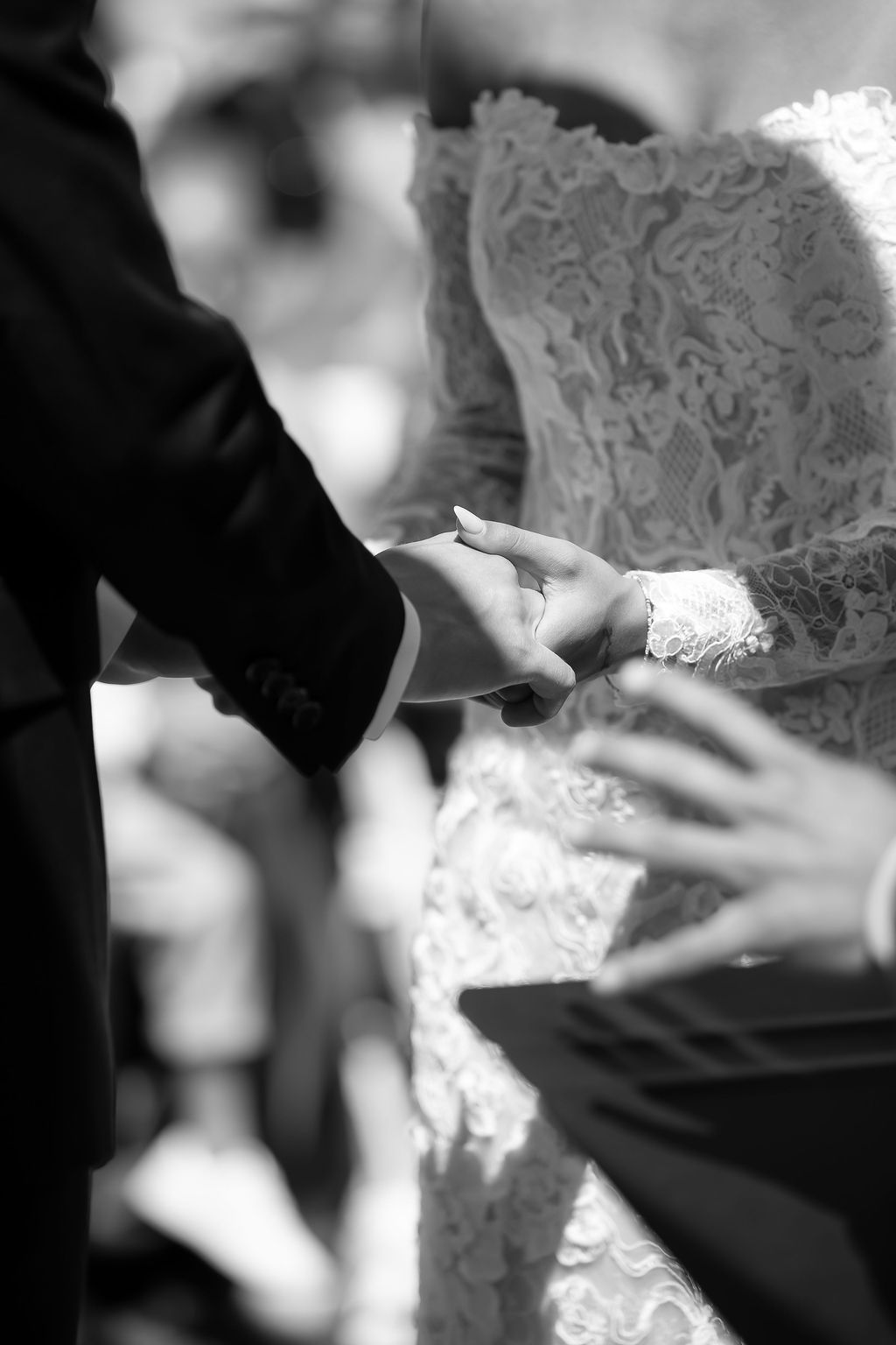 A black and white detail shot of hands clasped together, showing intricate lace wedding dress details.