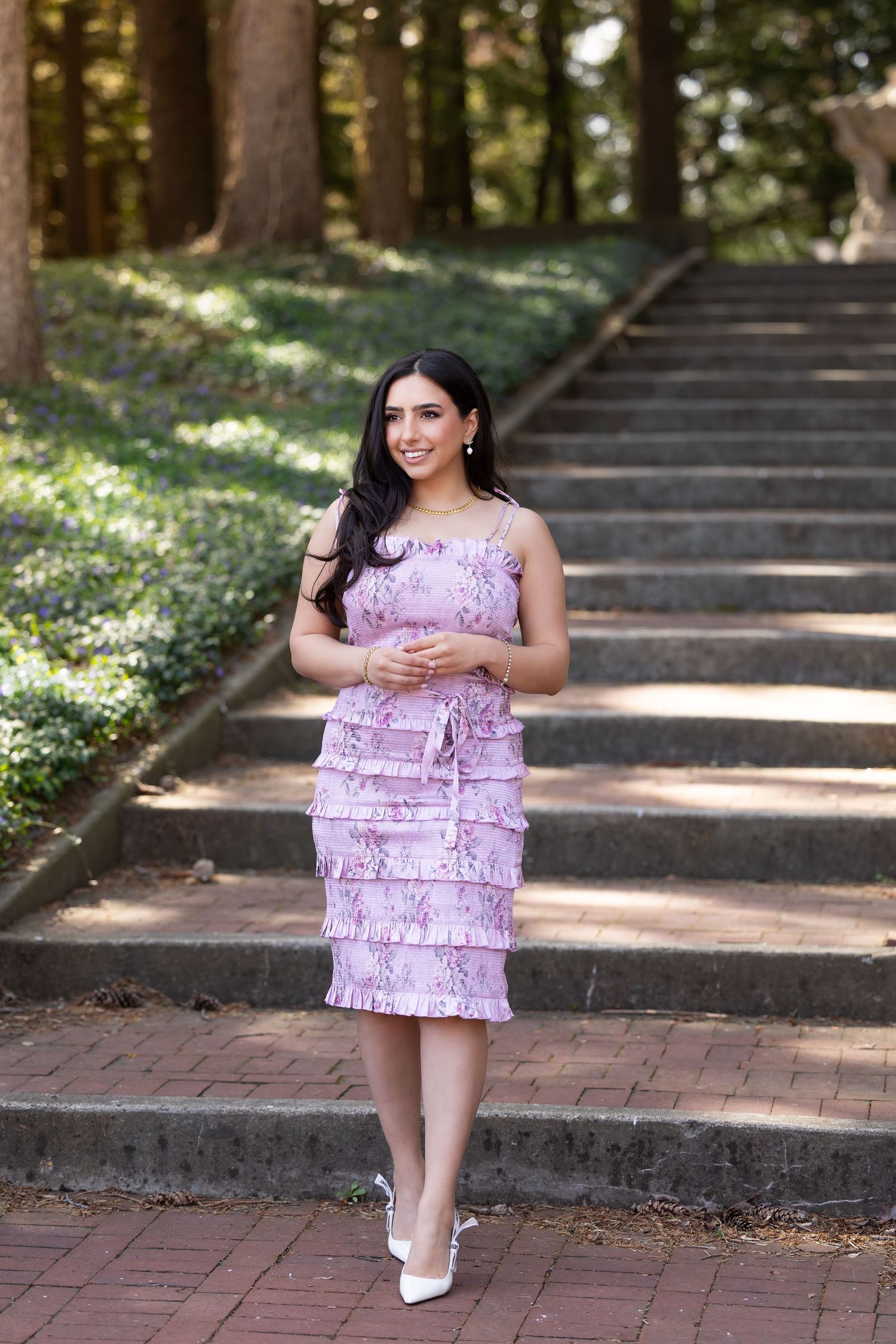 A woman in a pink tiered dress poses elegantly on brick stairs in a wooded garden setting.
