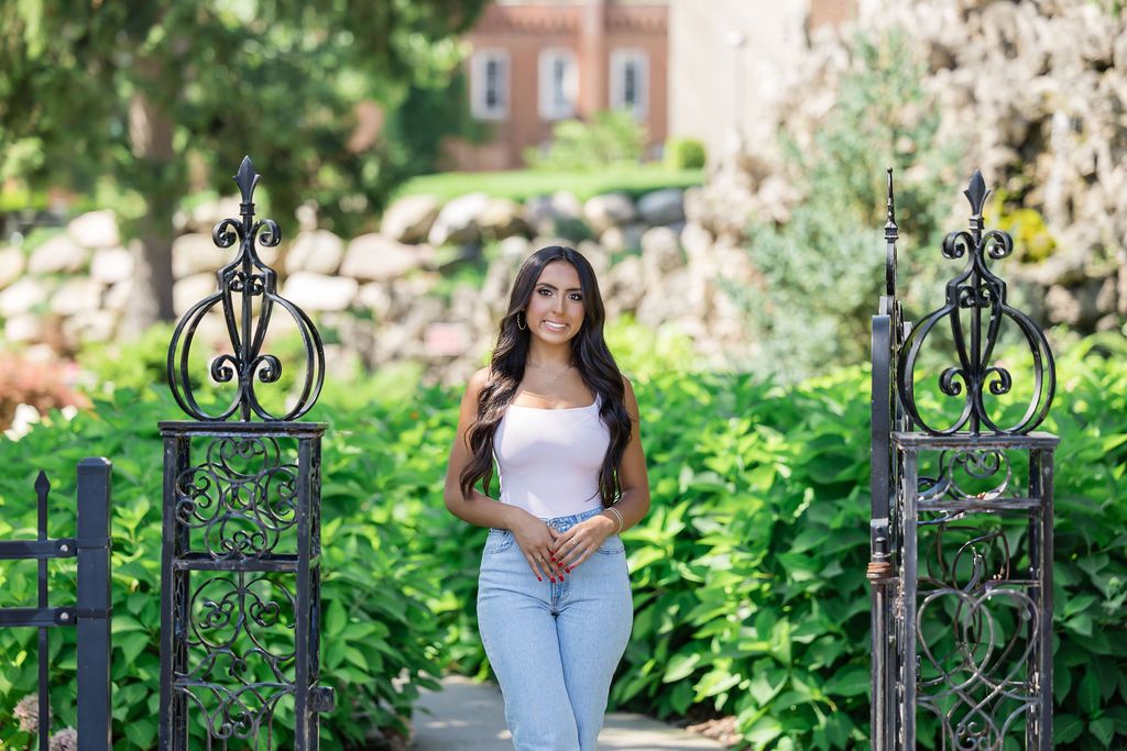 A decorative iron gate stands open to a lush garden path with greenery and a brick building in the background.