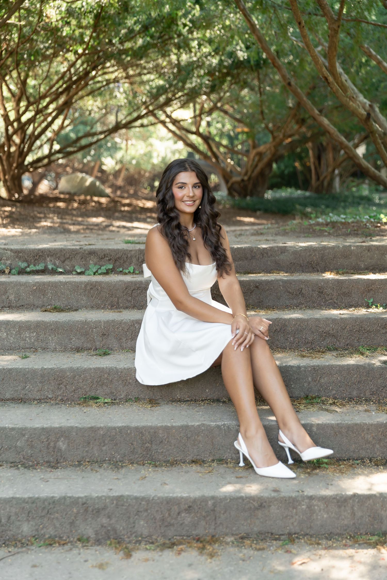 Woman in white dress and heels sits elegantly on outdoor stone steps.