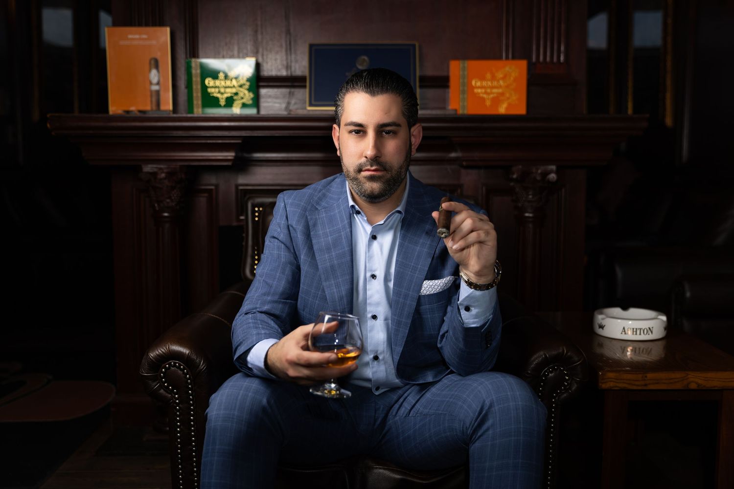 A gentleman in a blue suit sits in a leather chair holding a glass of whiskey in a dimly lit bar setting.