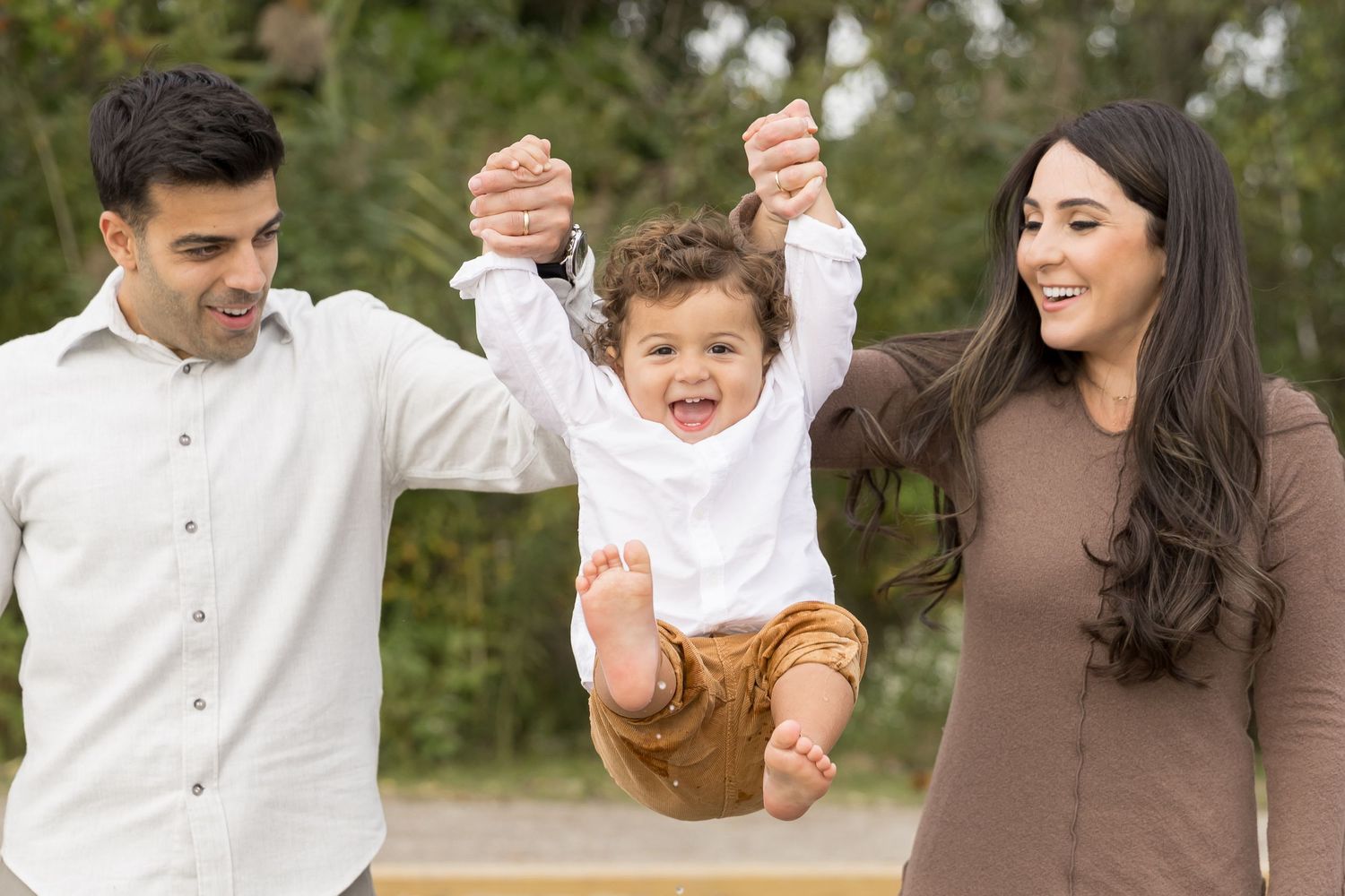 Parents playing outdoors and lifting joyful child in casual clothing among greenery on sunny day.