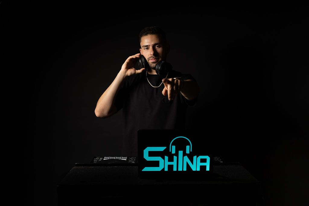 A DJ stands behind equipment with blue illuminated text reading SHINA on a dark background.
