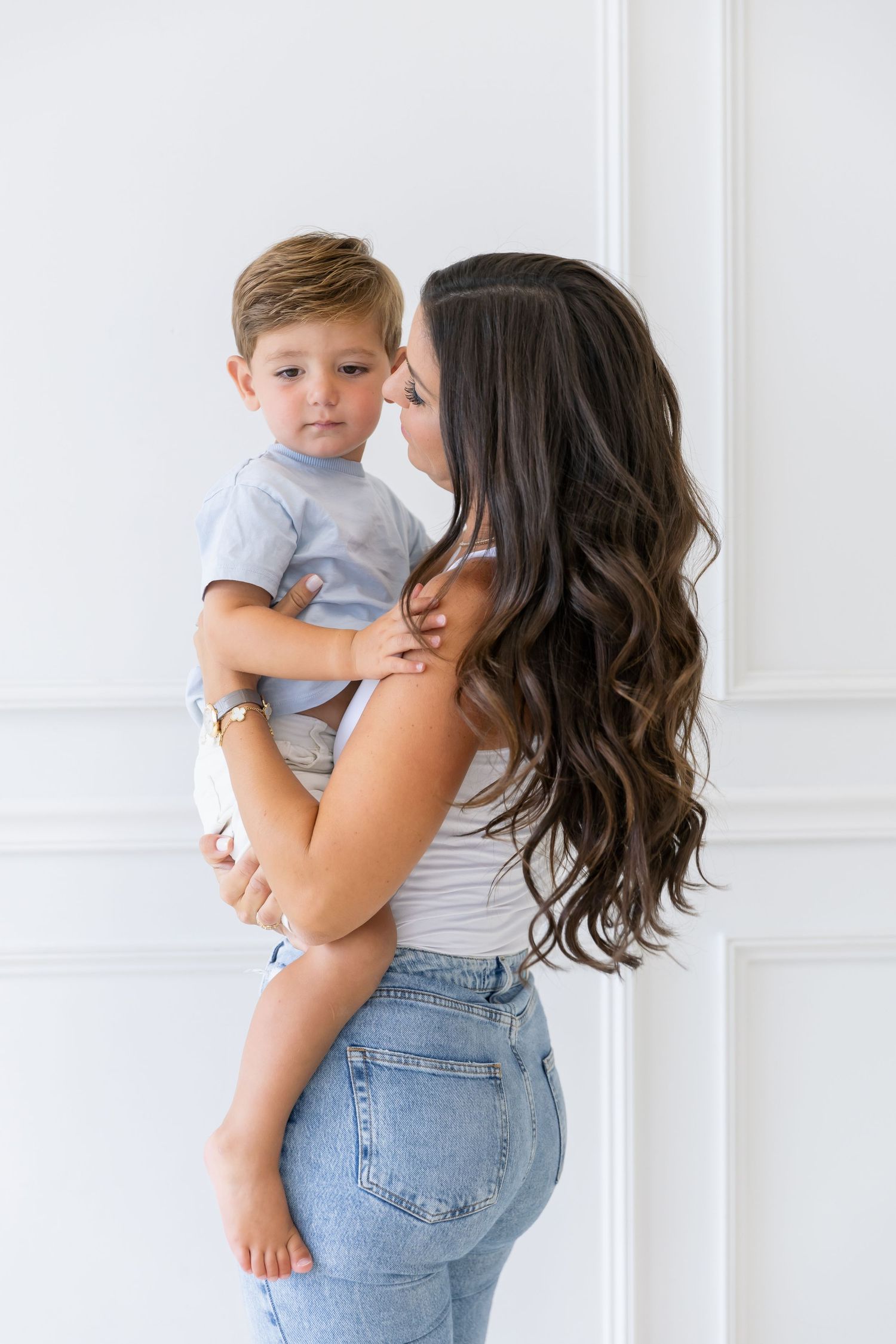 A parent in jeans and white tank top holds a small child wearing a gray t-shirt against a white wall.