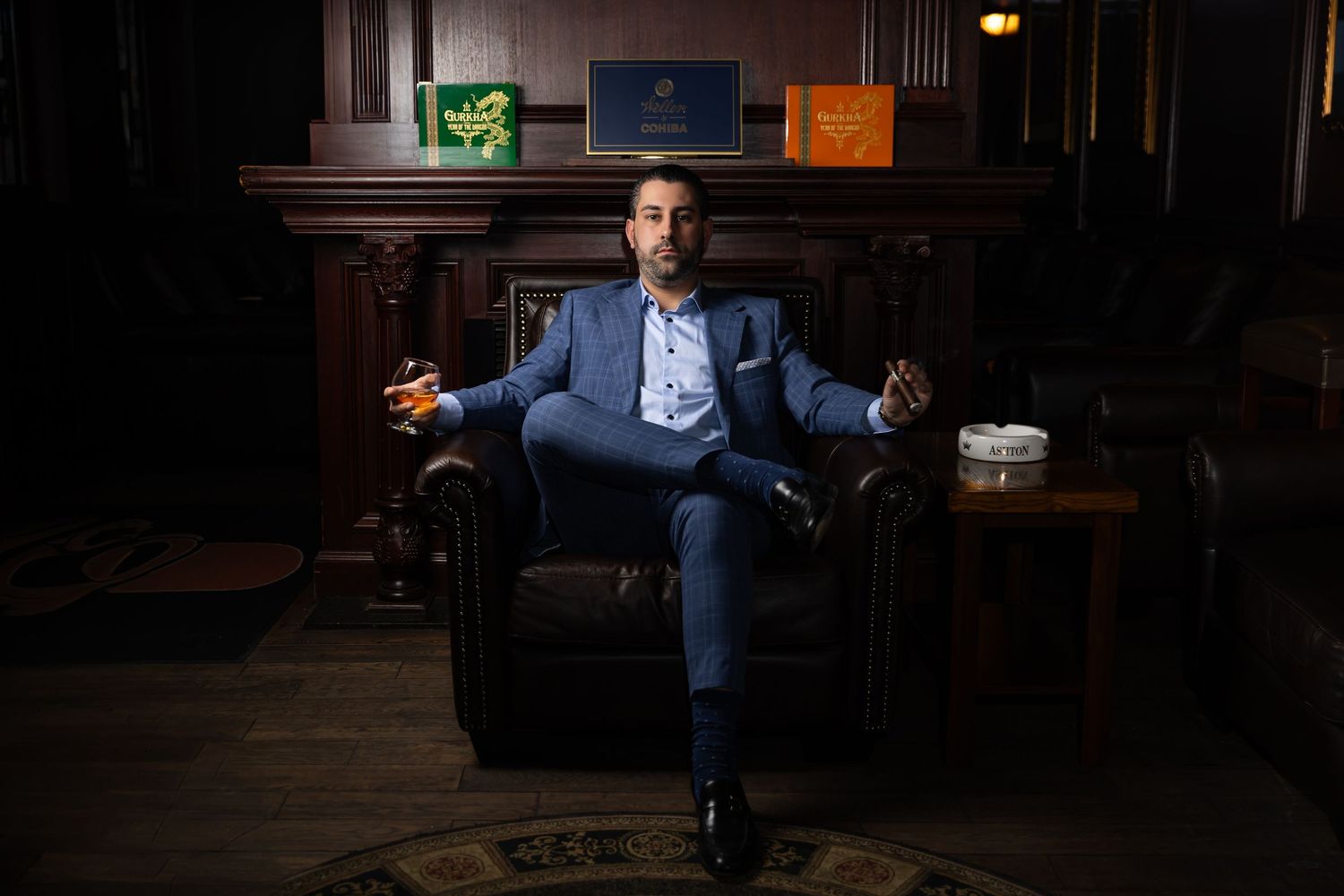 Person in blue suit sits confidently in leather armchair at dimly lit bar with drink in hand.