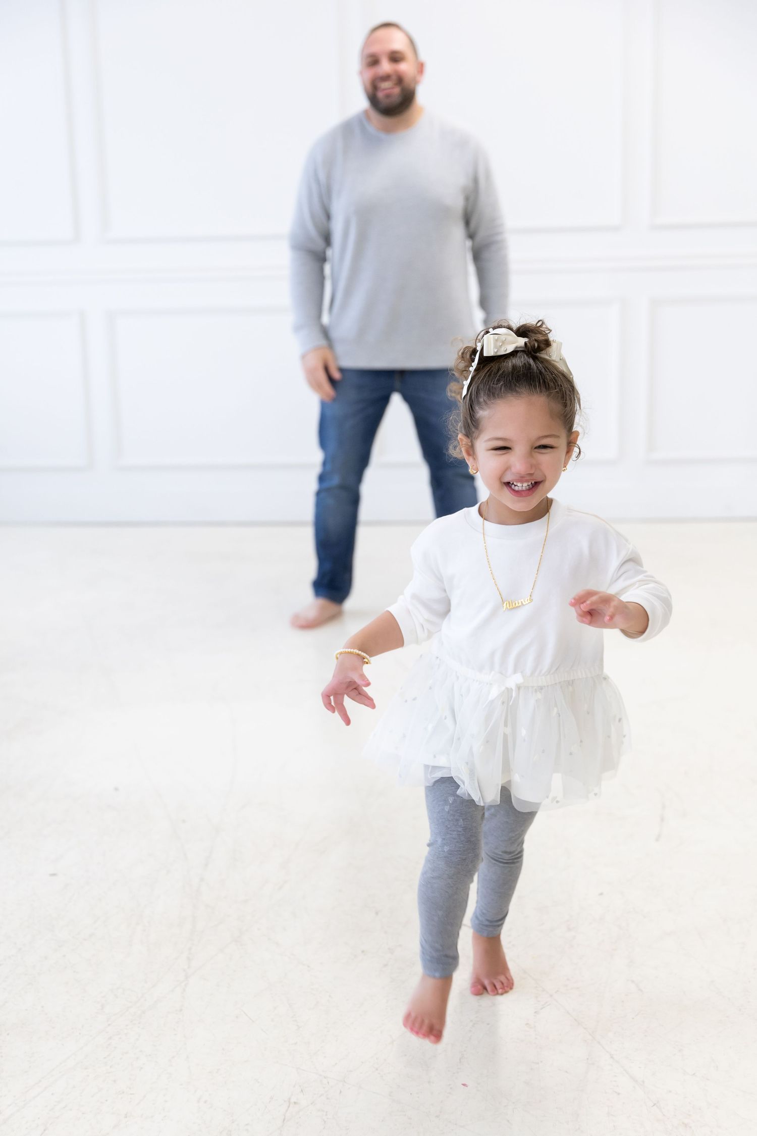 A child in a white dress and gray leggings dances barefoot on a light floor while someone watches from behind.