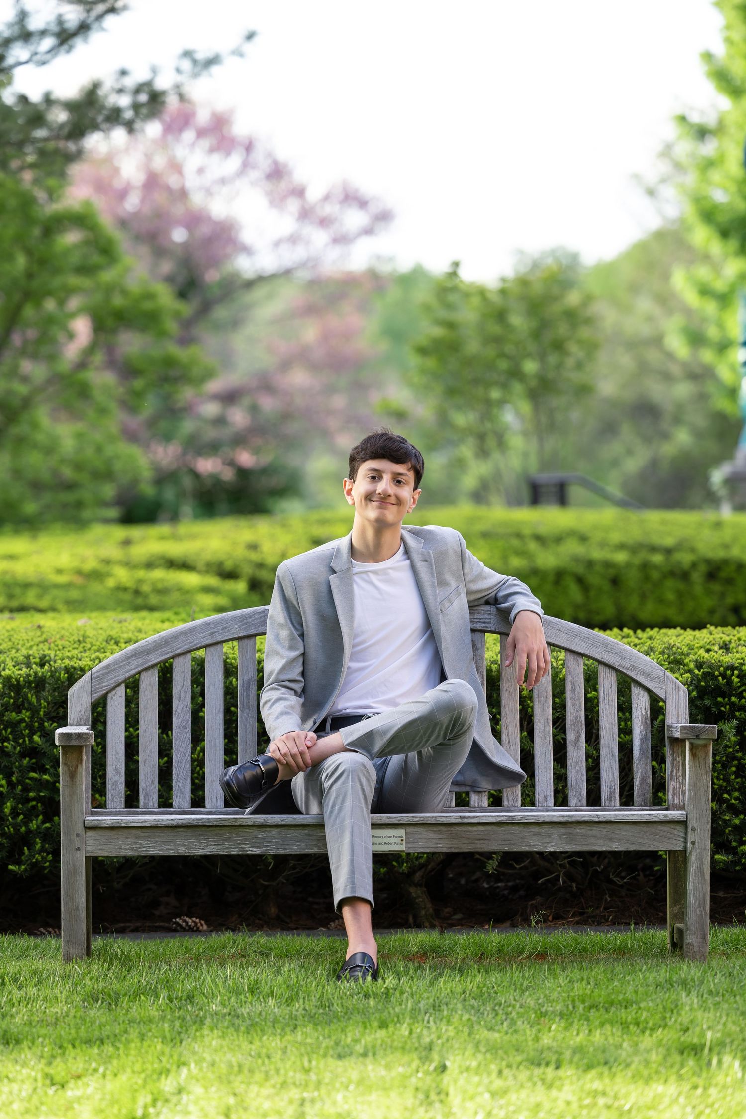 Person in grey suit sits casually on wooden bench in garden with blooming flowers.