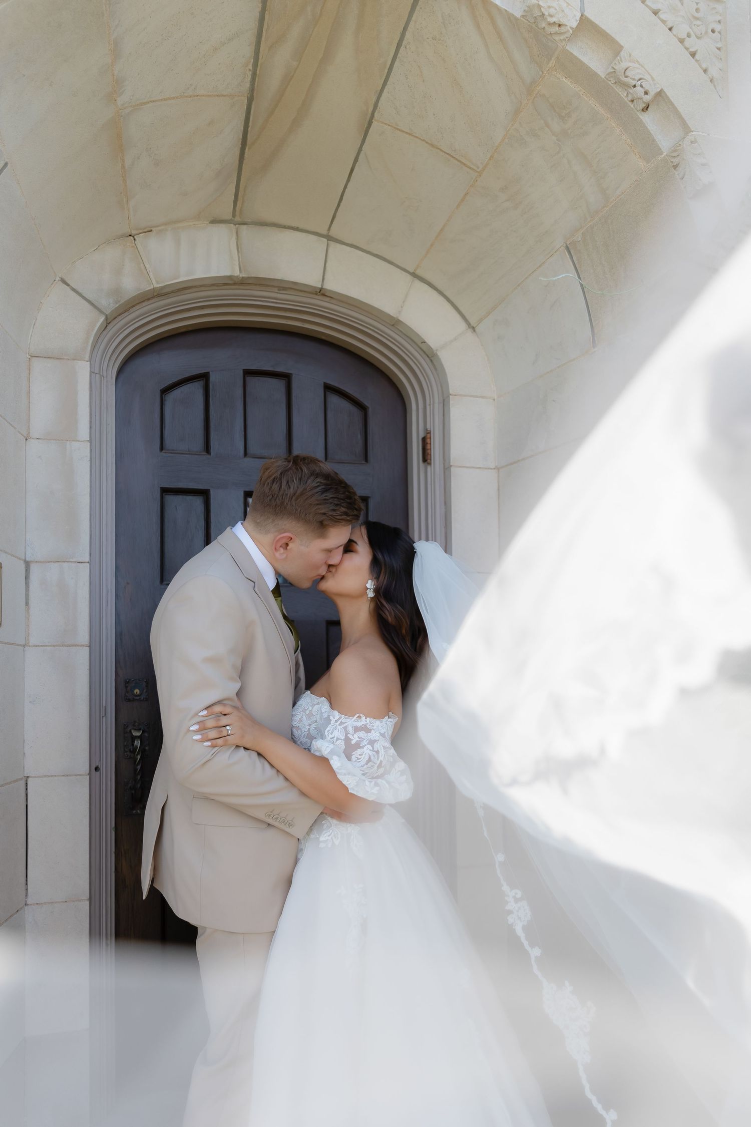 Newlyweds embrace in front of dark wooden door under stone archway.