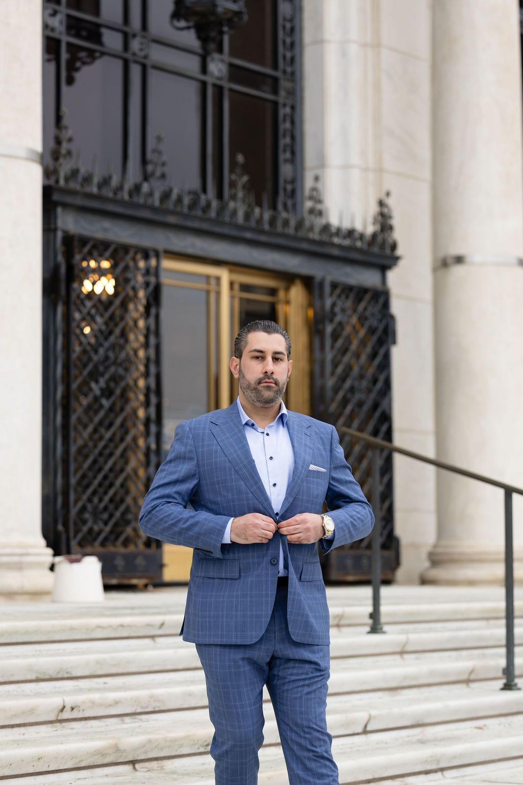 Professional wearing a blue checkered suit stands on marble steps in front of an ornate building entrance.