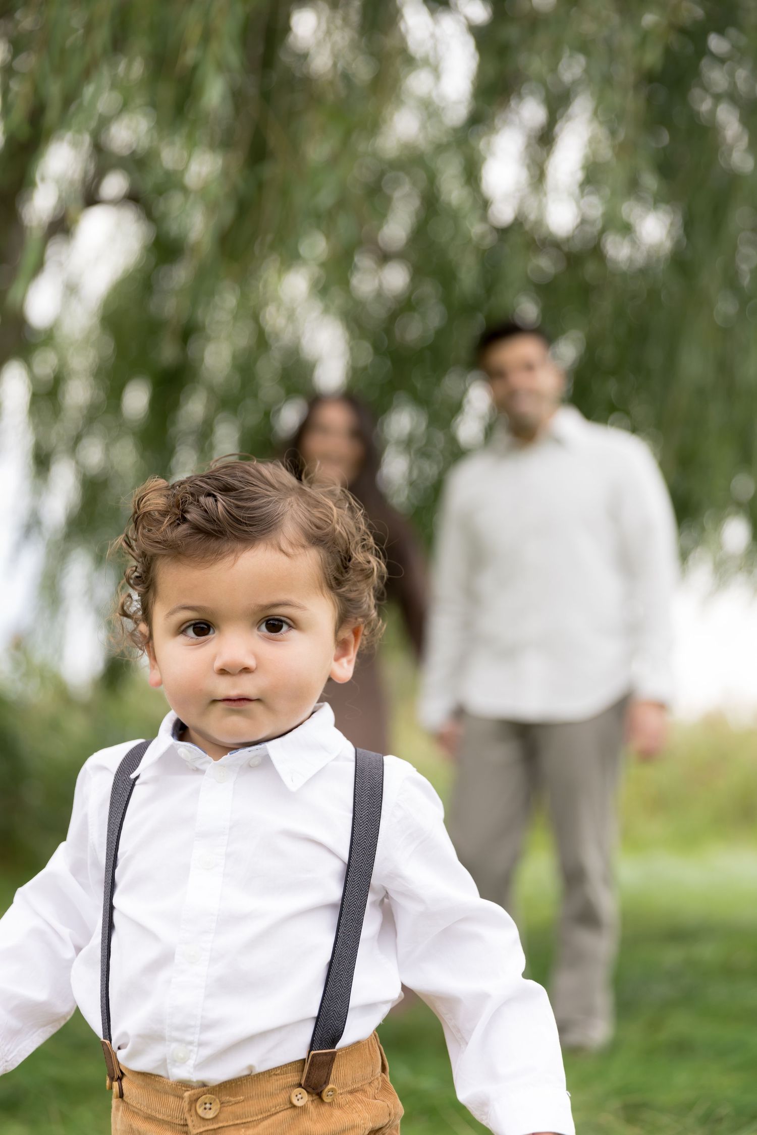 A young child in a white dress shirt and suspenders stands in a garden with blurred figures in the background.