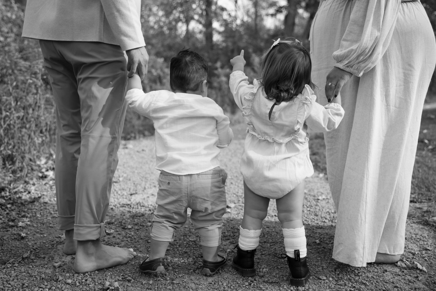 A black and white photograph shows two small toddlers holding hands while standing between adults outdoors.