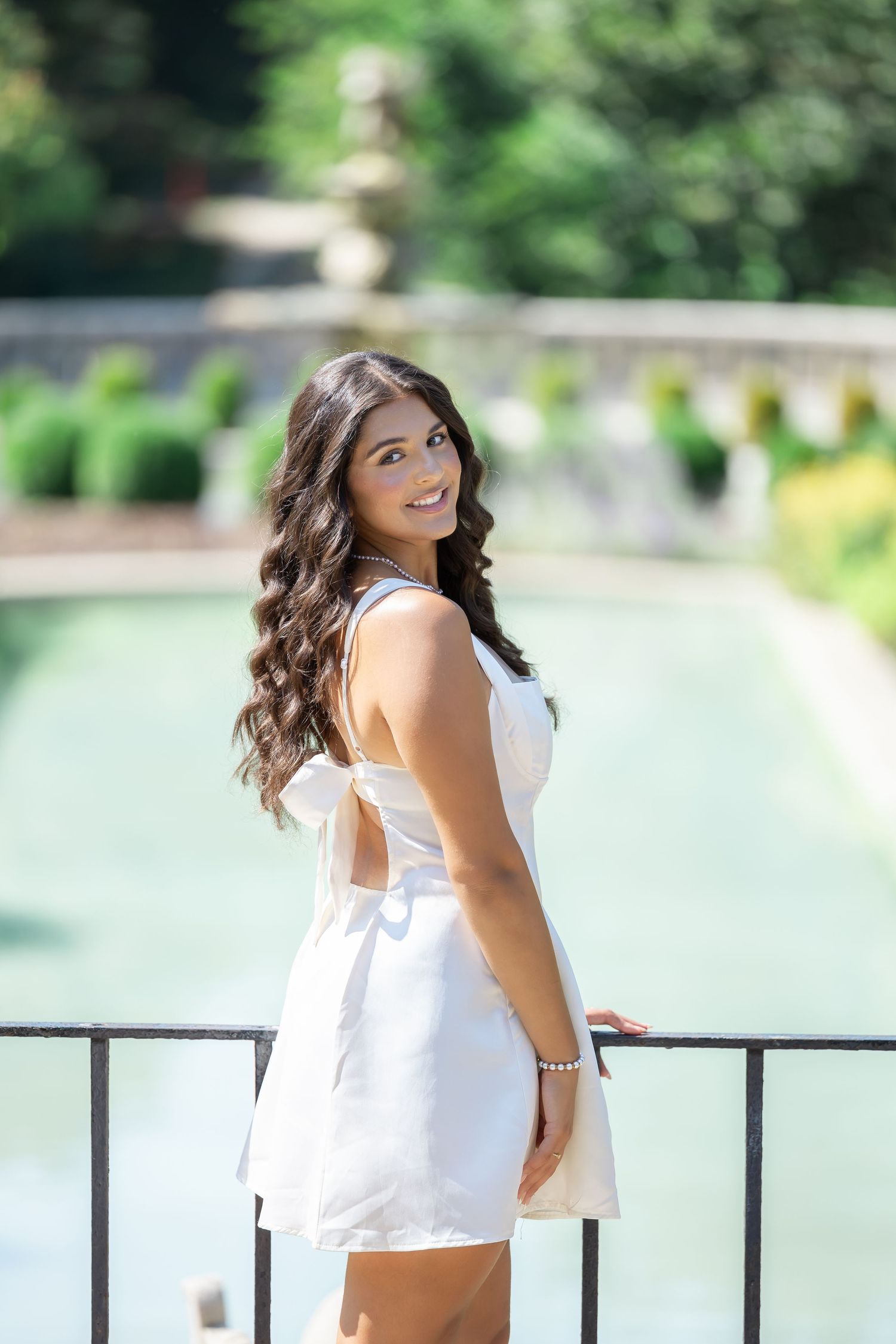 A woman in a white summer dress poses by a reflecting pool in a garden setting.