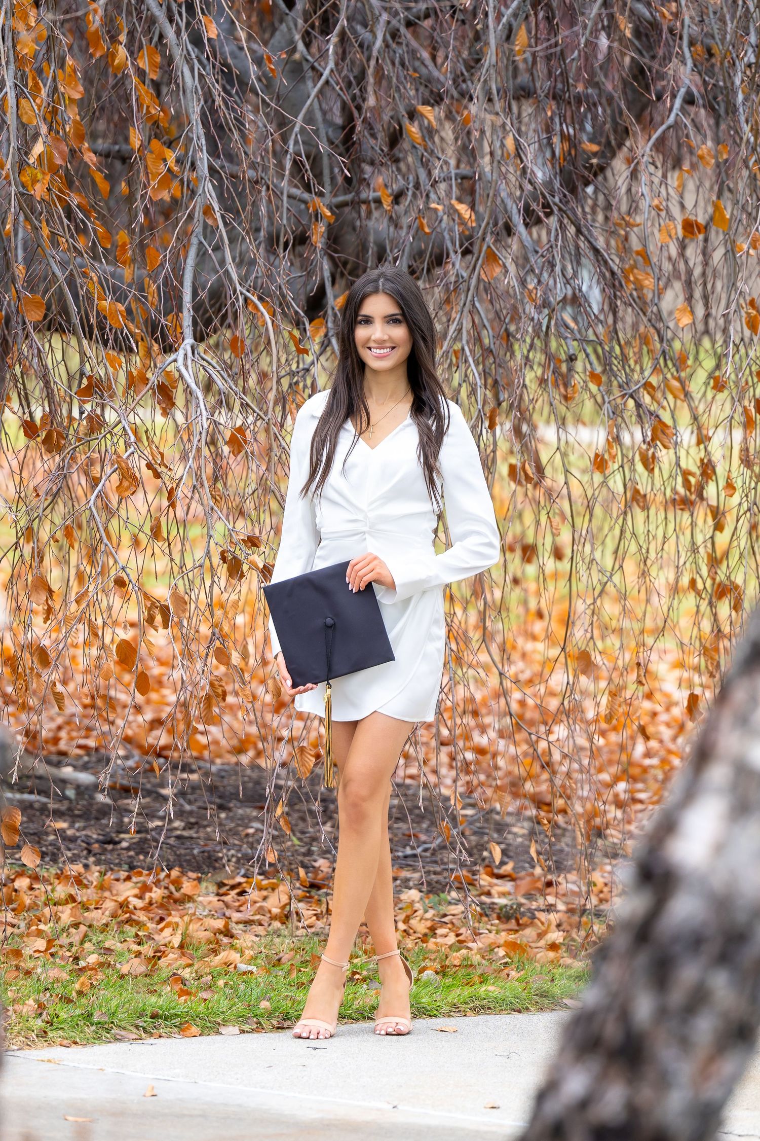 A woman in a white dress poses with graduation cap against an autumn nature backdrop.