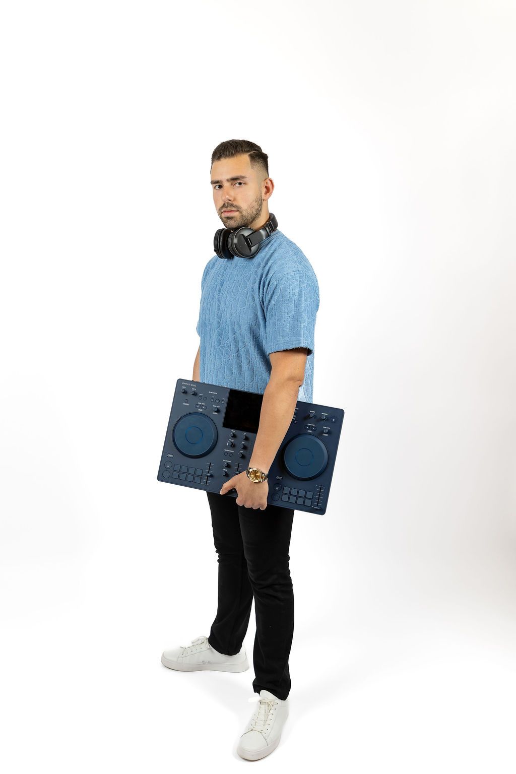 A DJ carrying professional turntable decks wearing a light blue polo shirt, dark jeans and white sneakers against white backdrop.