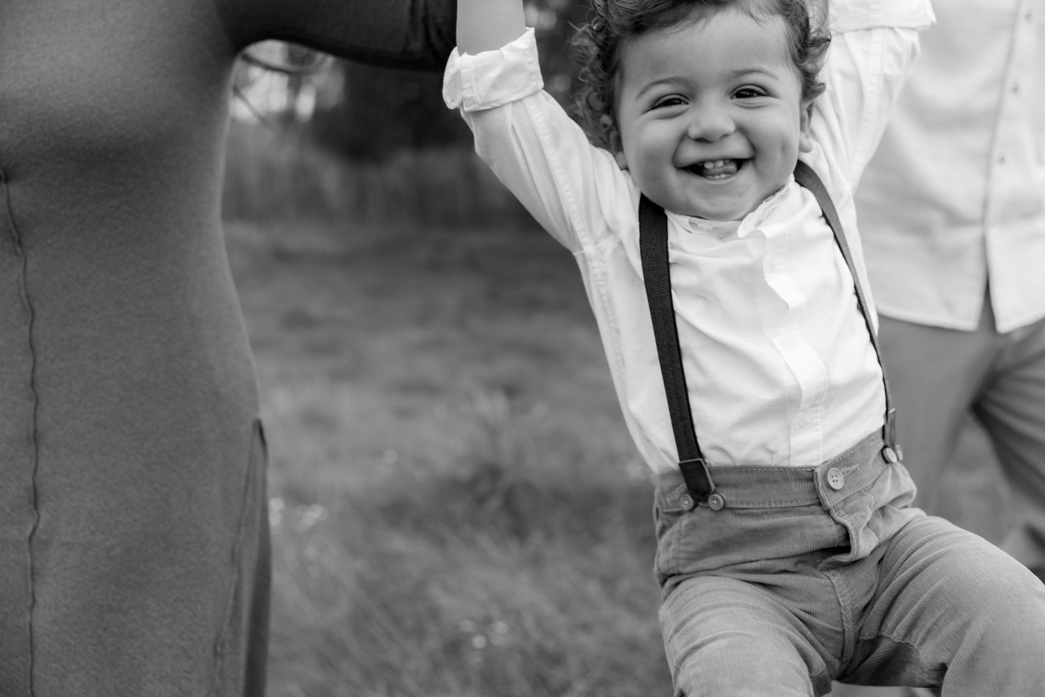 A joyful young child in suspenders and white shirt raises their arms while sitting in a black and white photo.