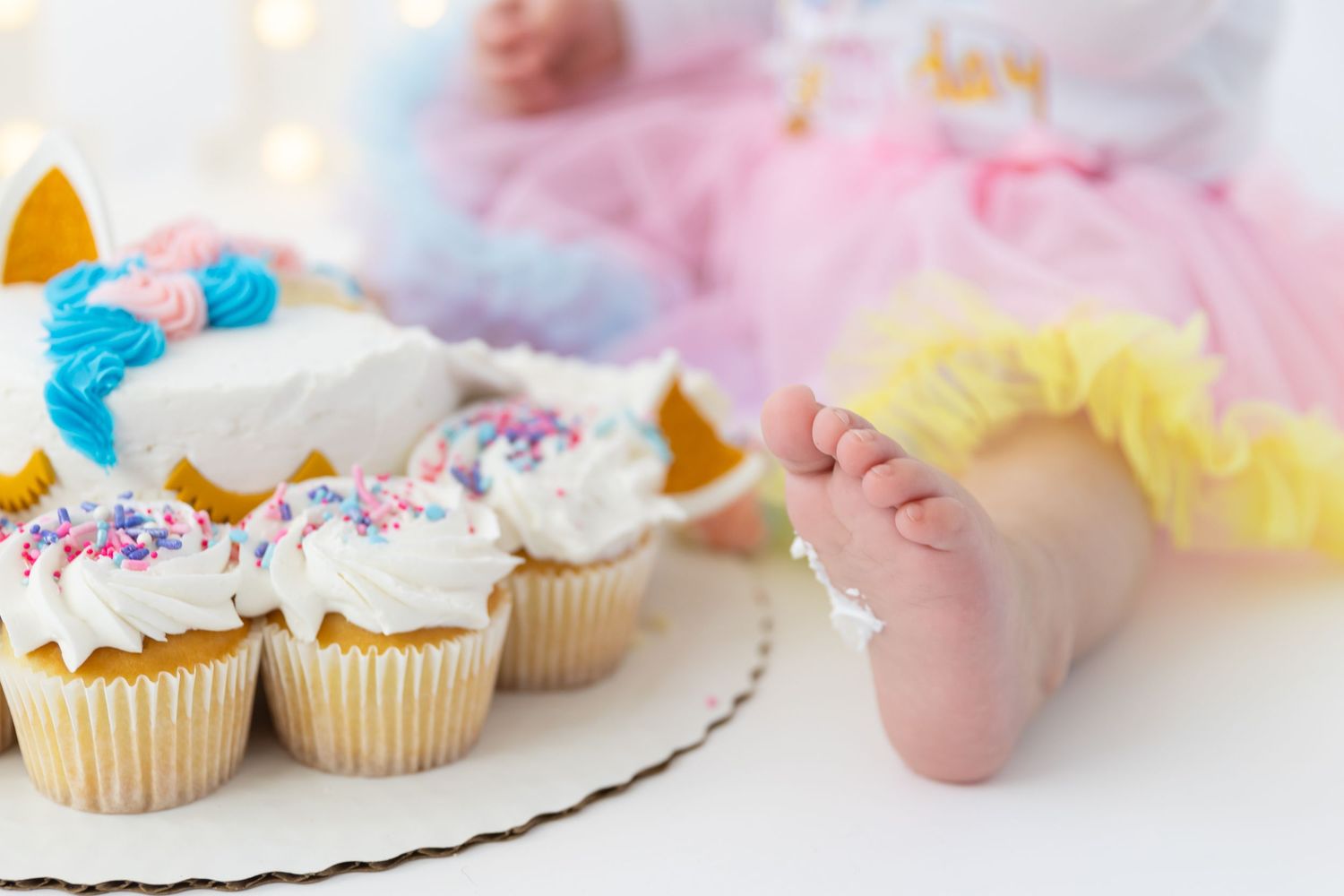 Decorated vanilla cupcakes with colorful sprinkles and frosting on a white plate next to small feet in a tutu.