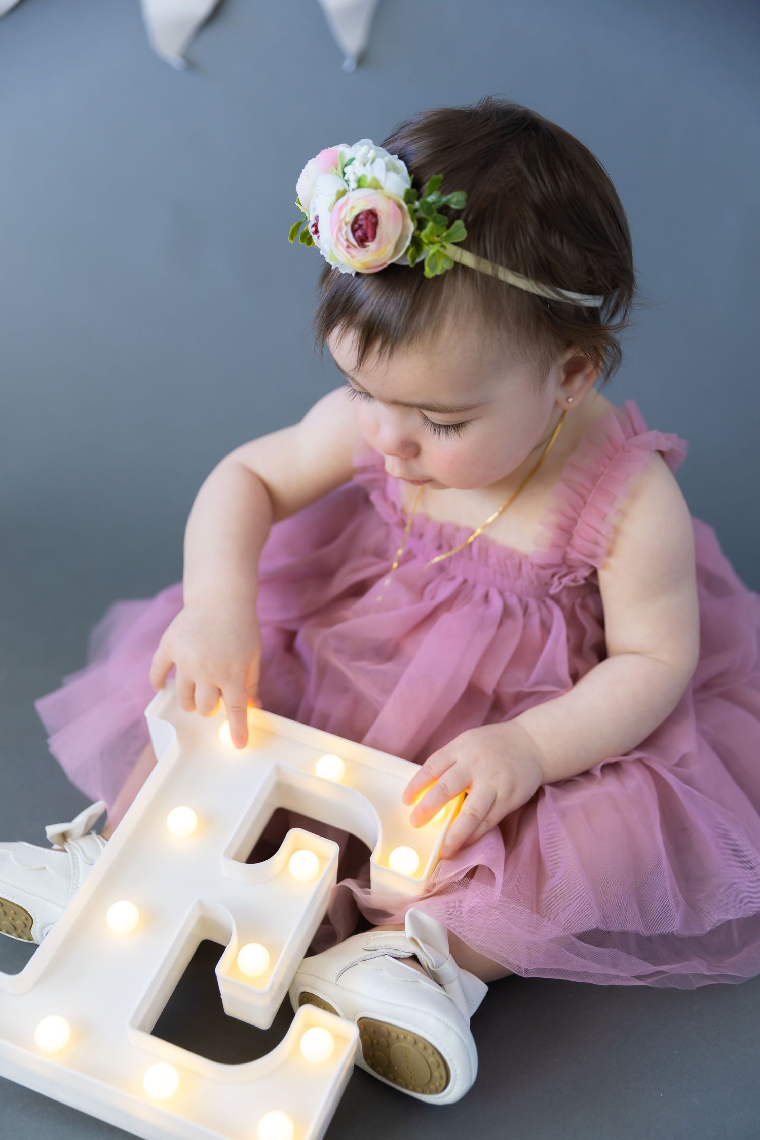 Baby in pink tulle dress touching illuminated letter E decoration while wearing floral headband on gray backdrop.