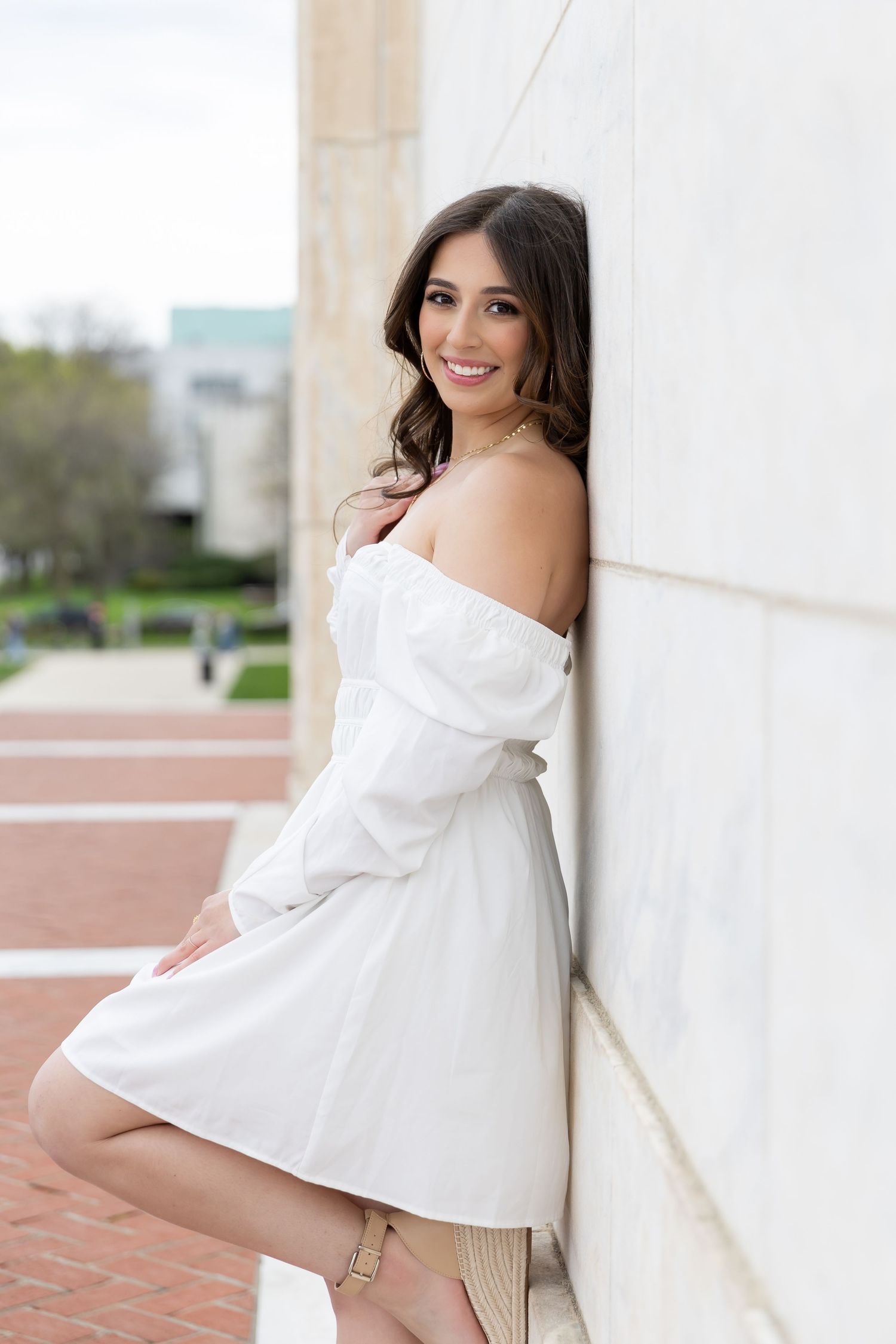 Woman in white off-shoulder dress poses against a white wall on a brick pathway.