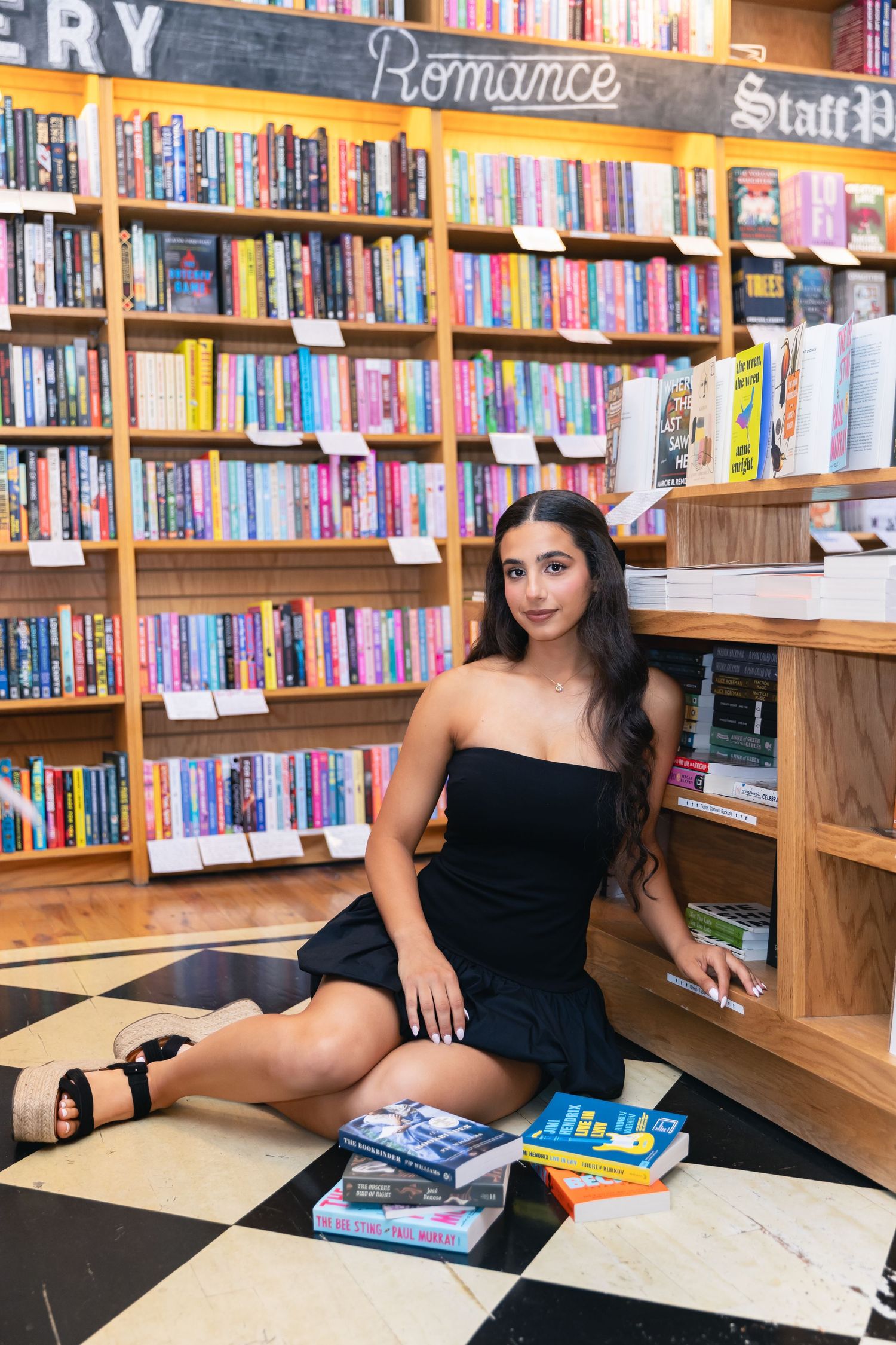 Young woman in black dress poses with books at a bookstore with colorful shelves in the background.