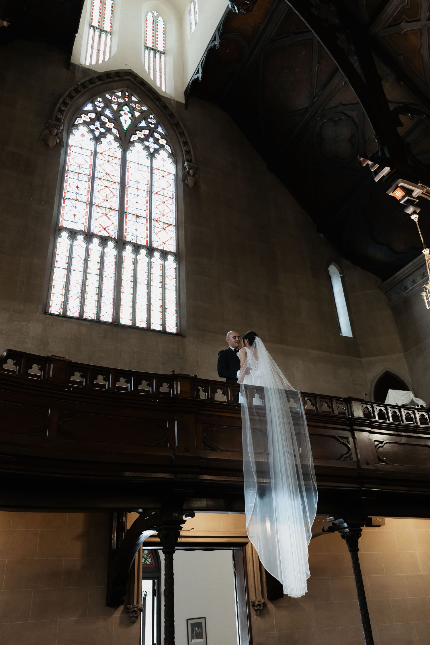 A dramatic chapel wedding photo featuring a gothic window and flowing white veil.