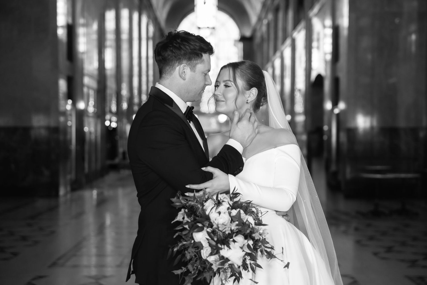 Romantic black and white wedding photo of couple embracing in elegant corridor with glowing lights.