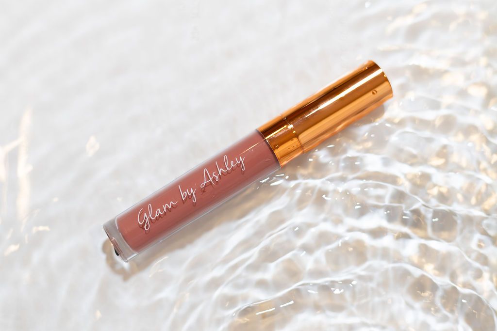 A rose gold liquid lipstick tube lying on a textured white surface with water droplet effects.