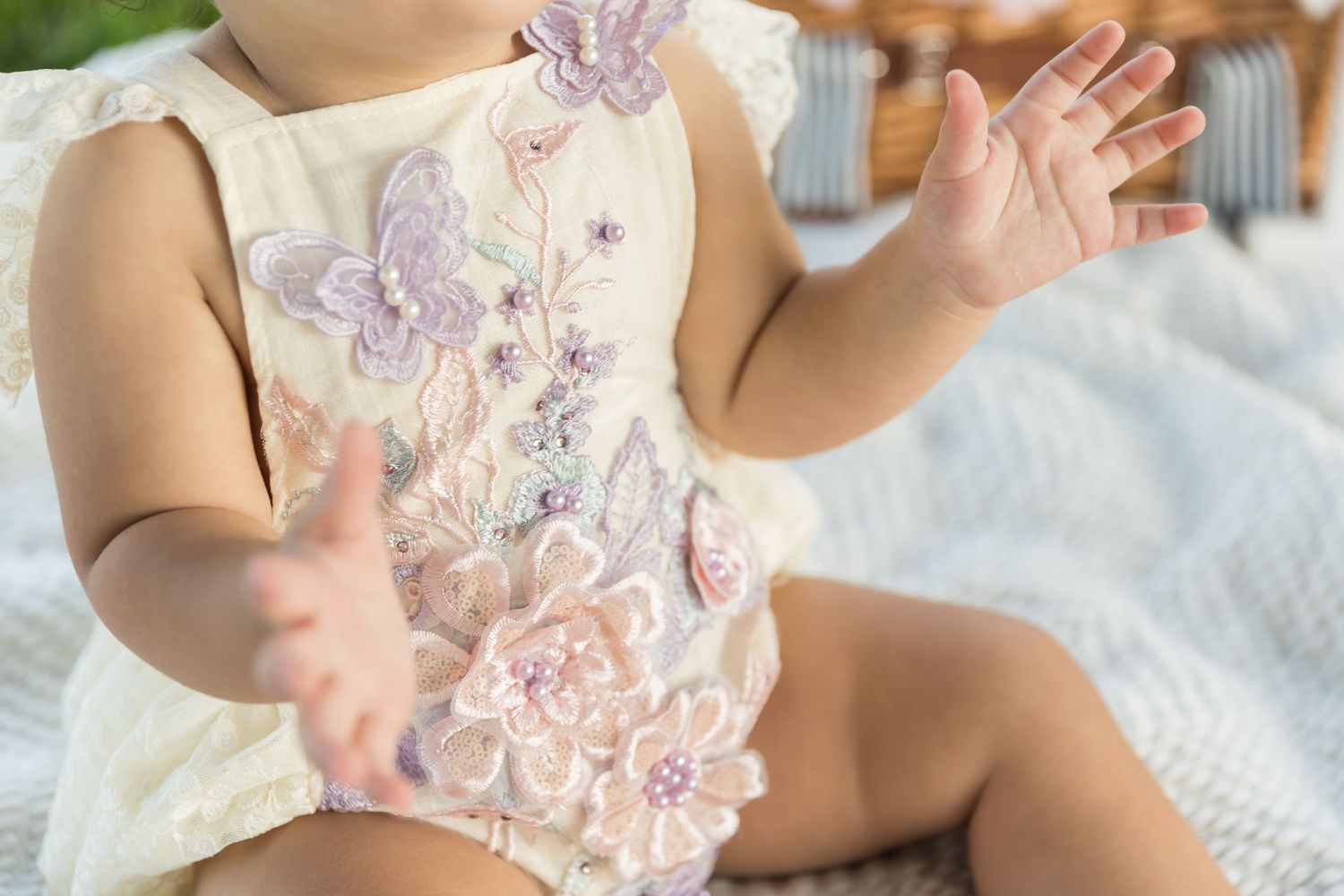 Baby wearing pastel floral romper with butterfly appliques sits on white blanket while reaching out with small hands.
