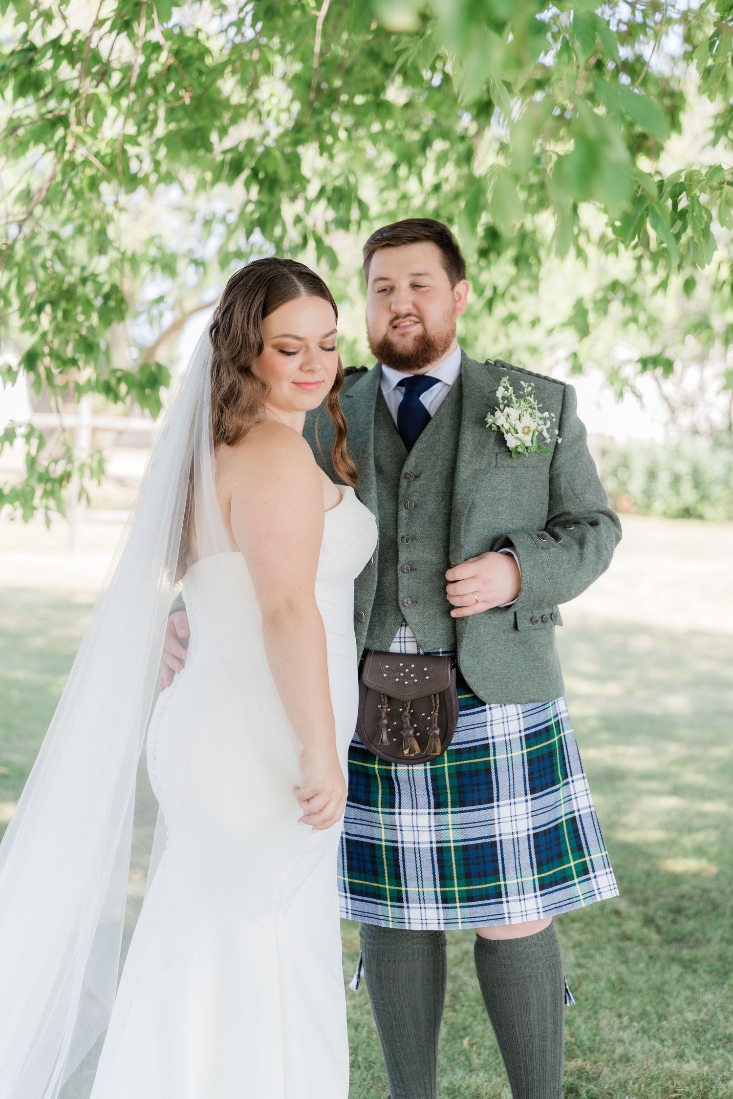 Bride in white gown and groom in traditional Scottish kilt standing together under blooming trees.