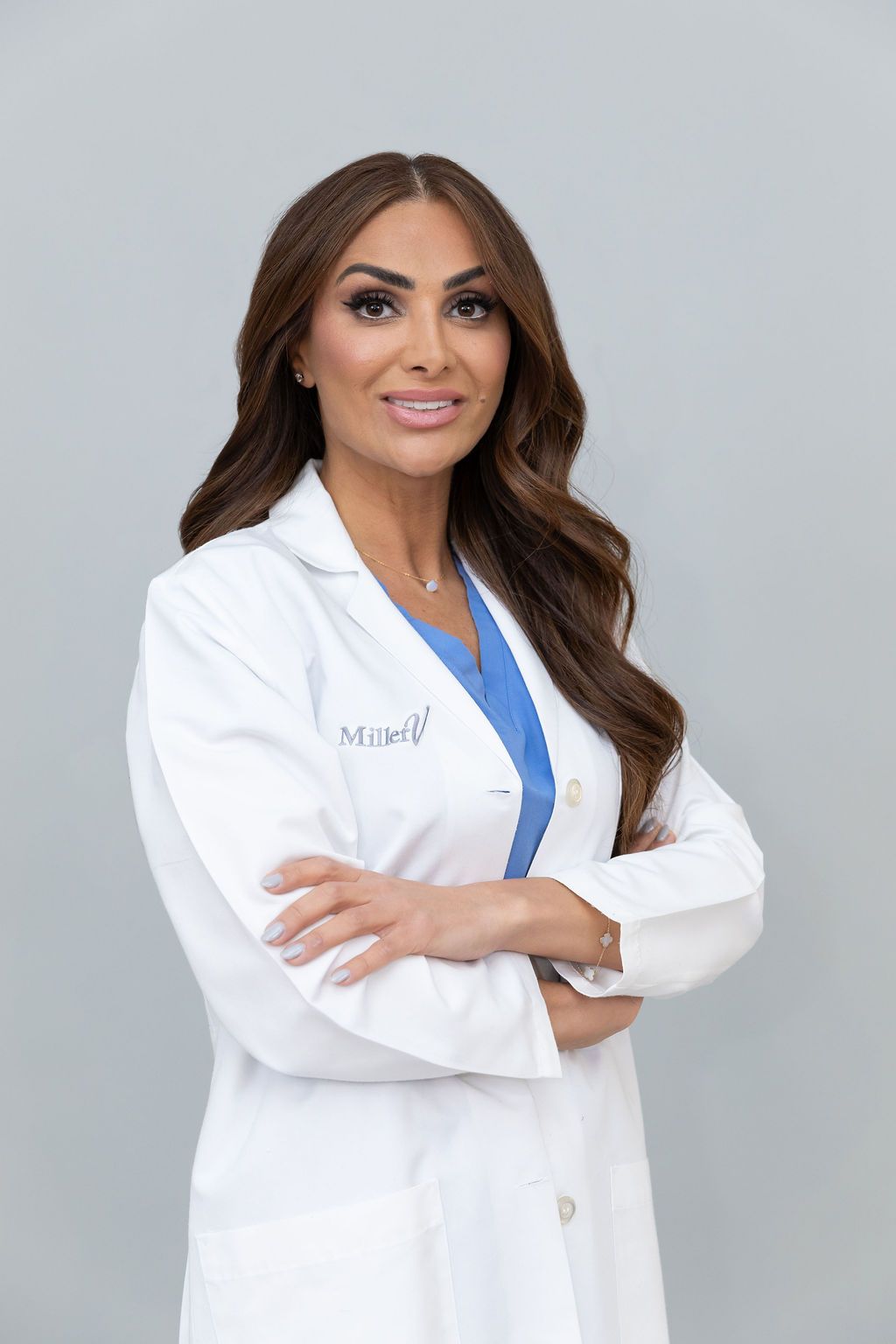 Medical professional in white lab coat and blue scrubs smiling confidently against gray background.