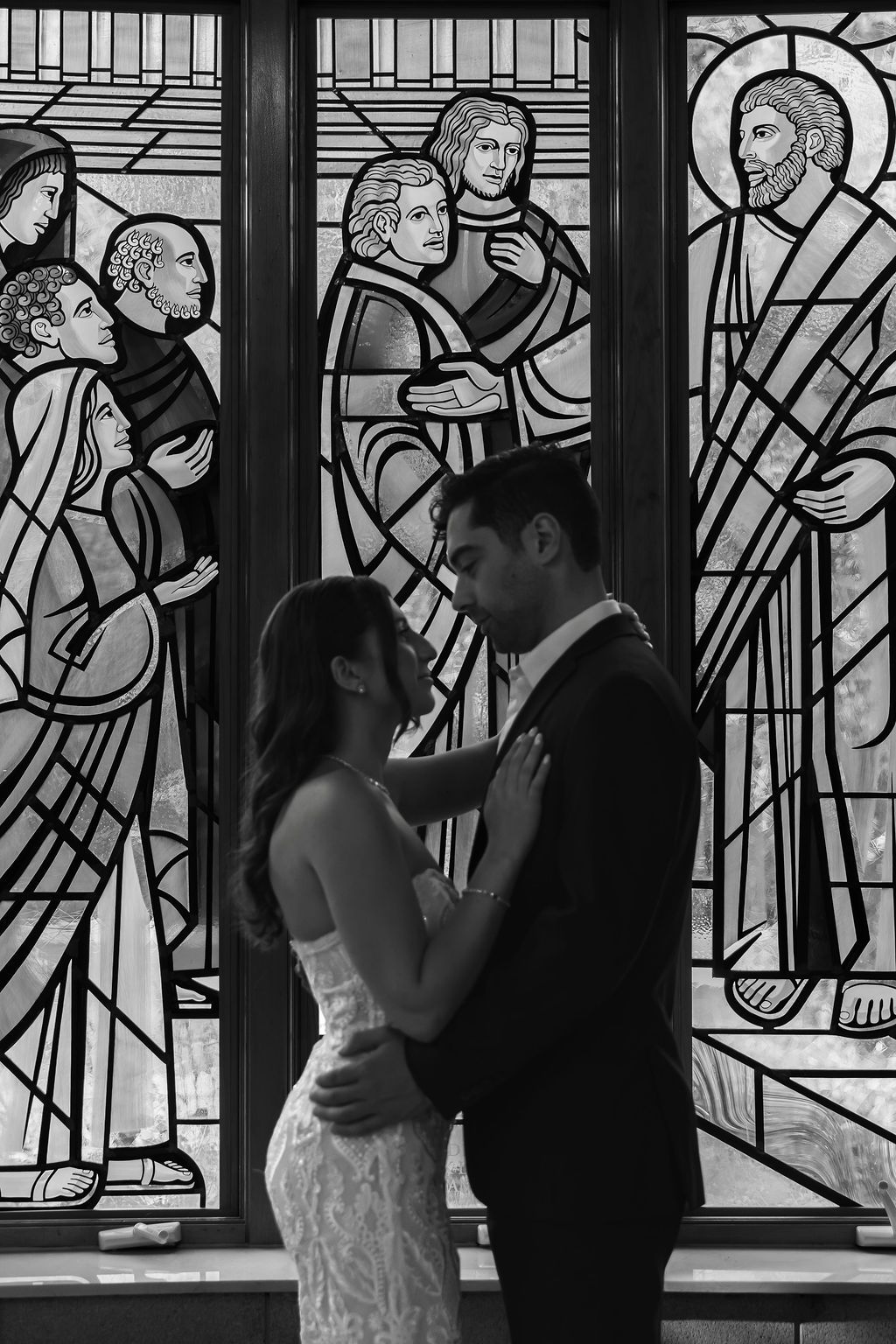 Silhouetted couple poses against dramatic religious stained glass windows in black and white.