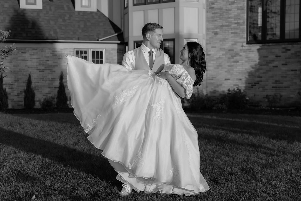 A romantic black and white photo of newlyweds dancing, with the bride's dress flowing dramatically against a stone building.