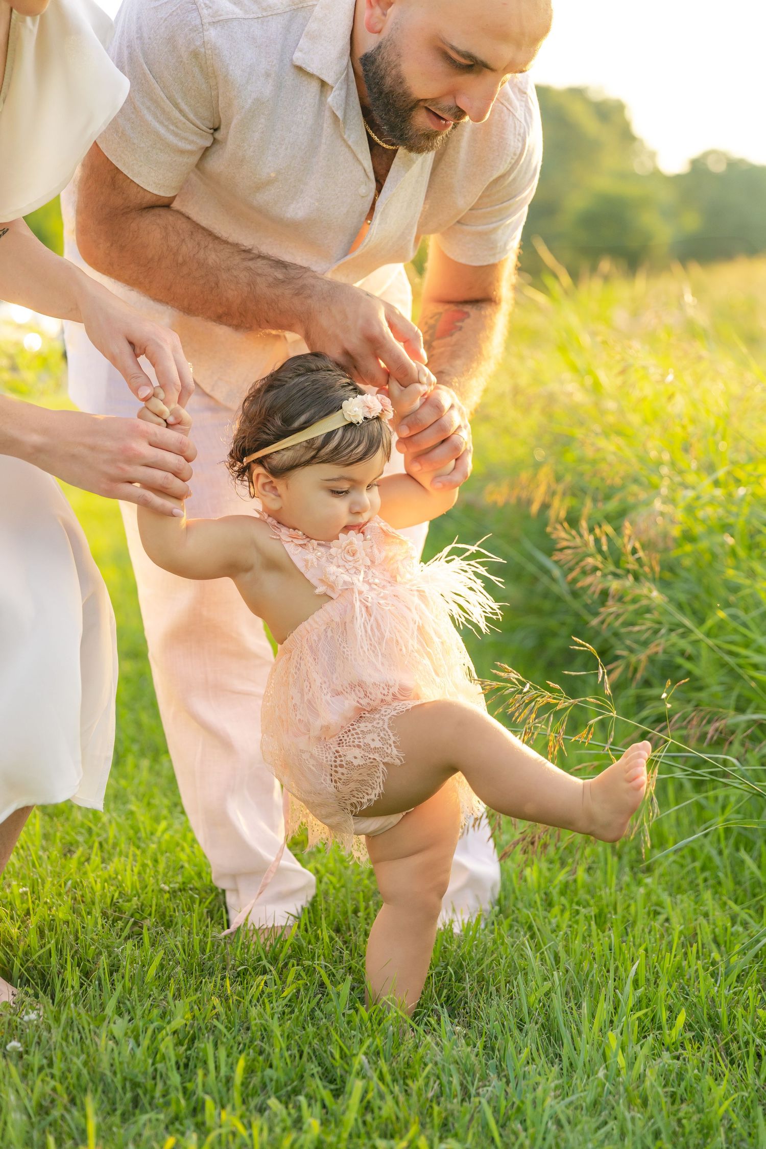 A parent helps a toddler in a lace dress take first steps on grass during golden hour sunset light.
