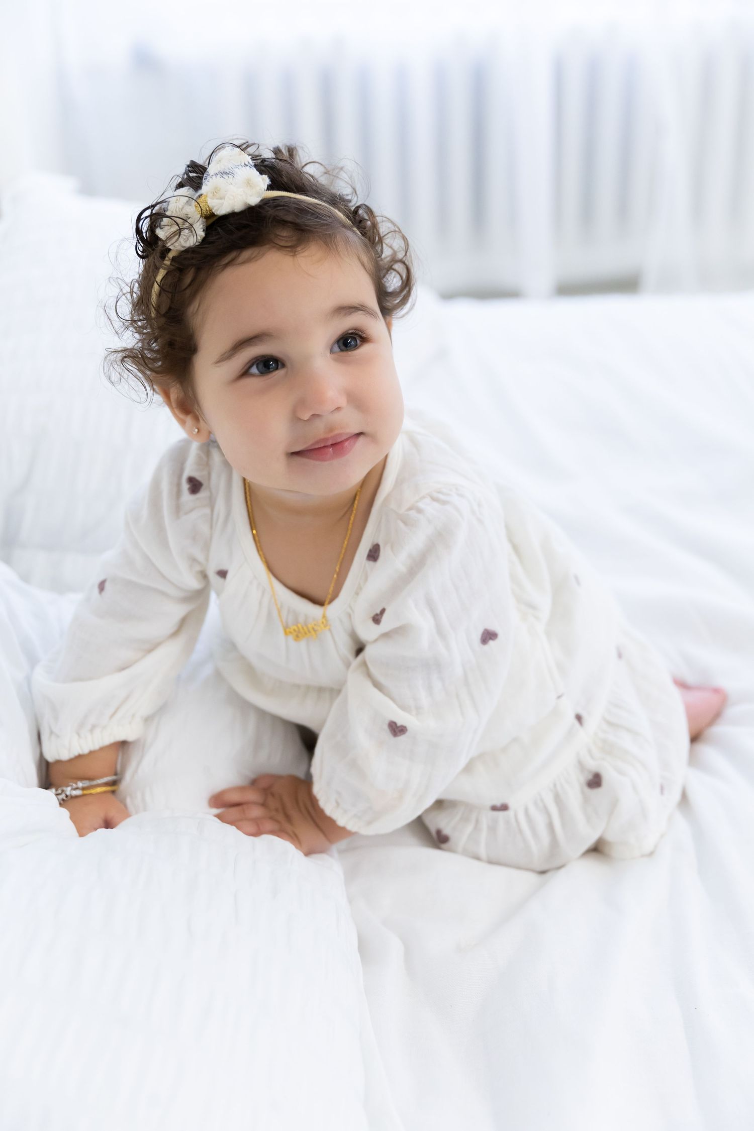 Child in white knitted outfit with floral headband lying on white bedding in bright indoor setting.